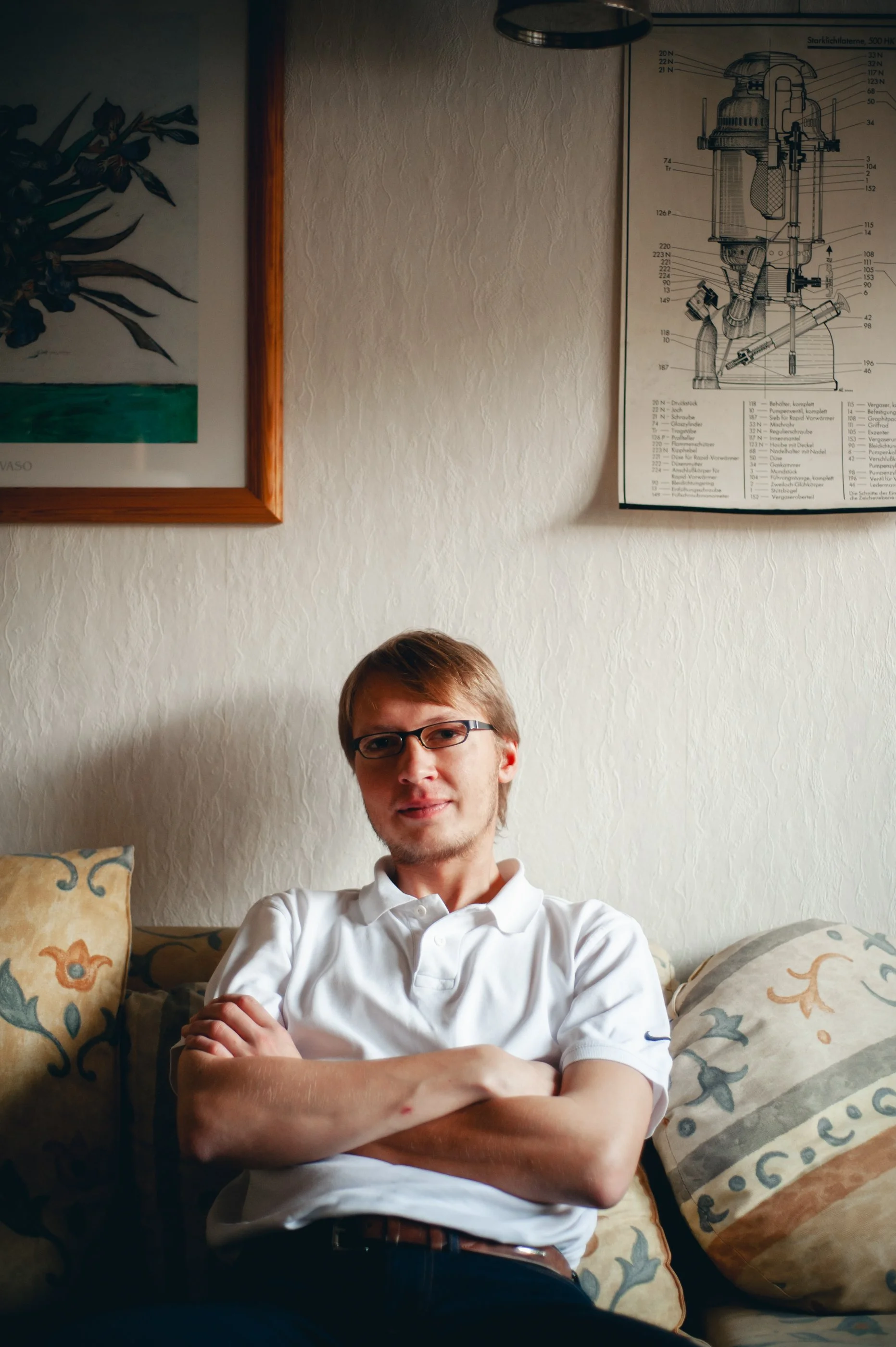 Ein junger Mann mit Brille sitzt auf einem Sofa mit kreisförmigen Mustern und posiert mit verschränkten Armen, im Hintergrund eine weiße Wand mit zwei Bildern, eines zeigt eine Illustration, das andere eine technische Zeichnung.