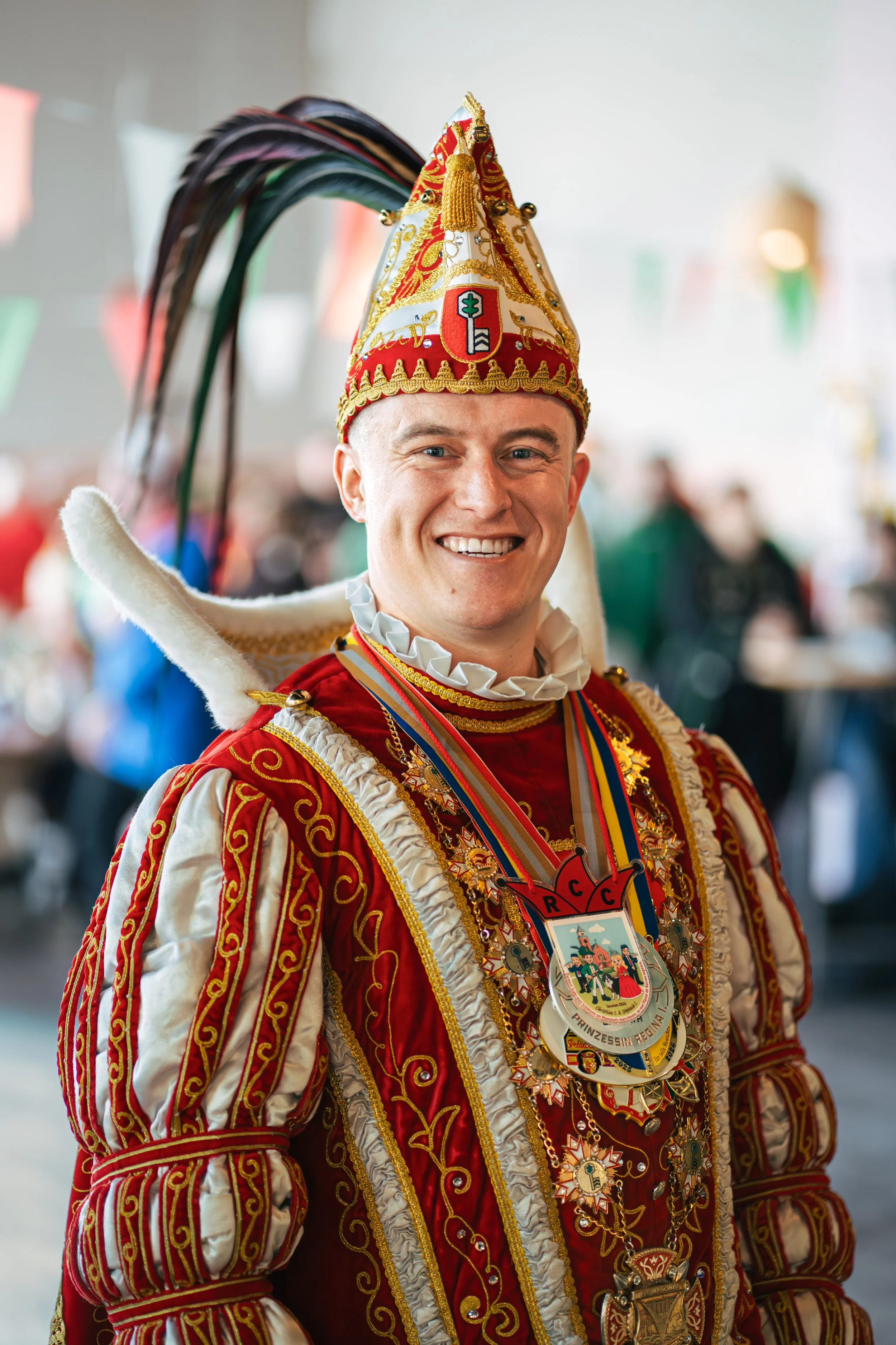 Ein Mann in einem festlichen, roten und goldenen Kostüm, trägt einen Zylinder mit bunten Federn und eine Vielzahl von Orden und Medaillen, während er lächelt.