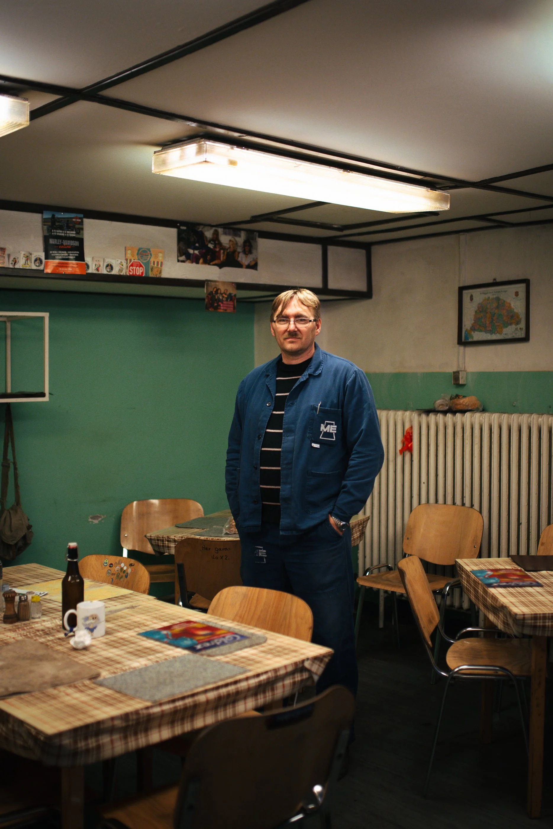 Ein Mann steht in einem rustikalen Restaurant mit Holztischen, Stühlen und einem grünen Wandabschnitt im Hintergrund. Es gibt einen Radiator an der Wand, sowie einige Fotos und Poster an der Decke hängen.