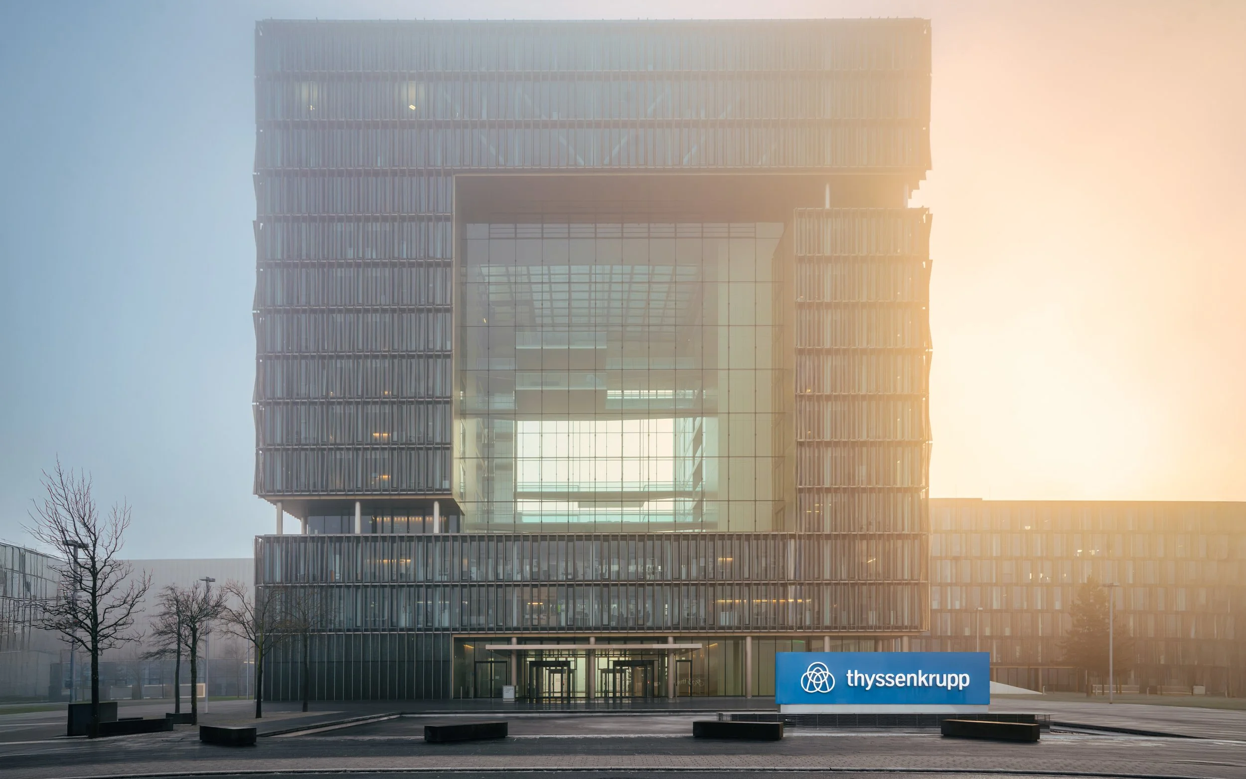 Modernes Hochhausgebäude mit Glasfassade und geöffnetem Innenraum, Sonnenaufgang im Hintergrund, ThyssenKrupp Logo auf einem Schild vor dem Gebäude.