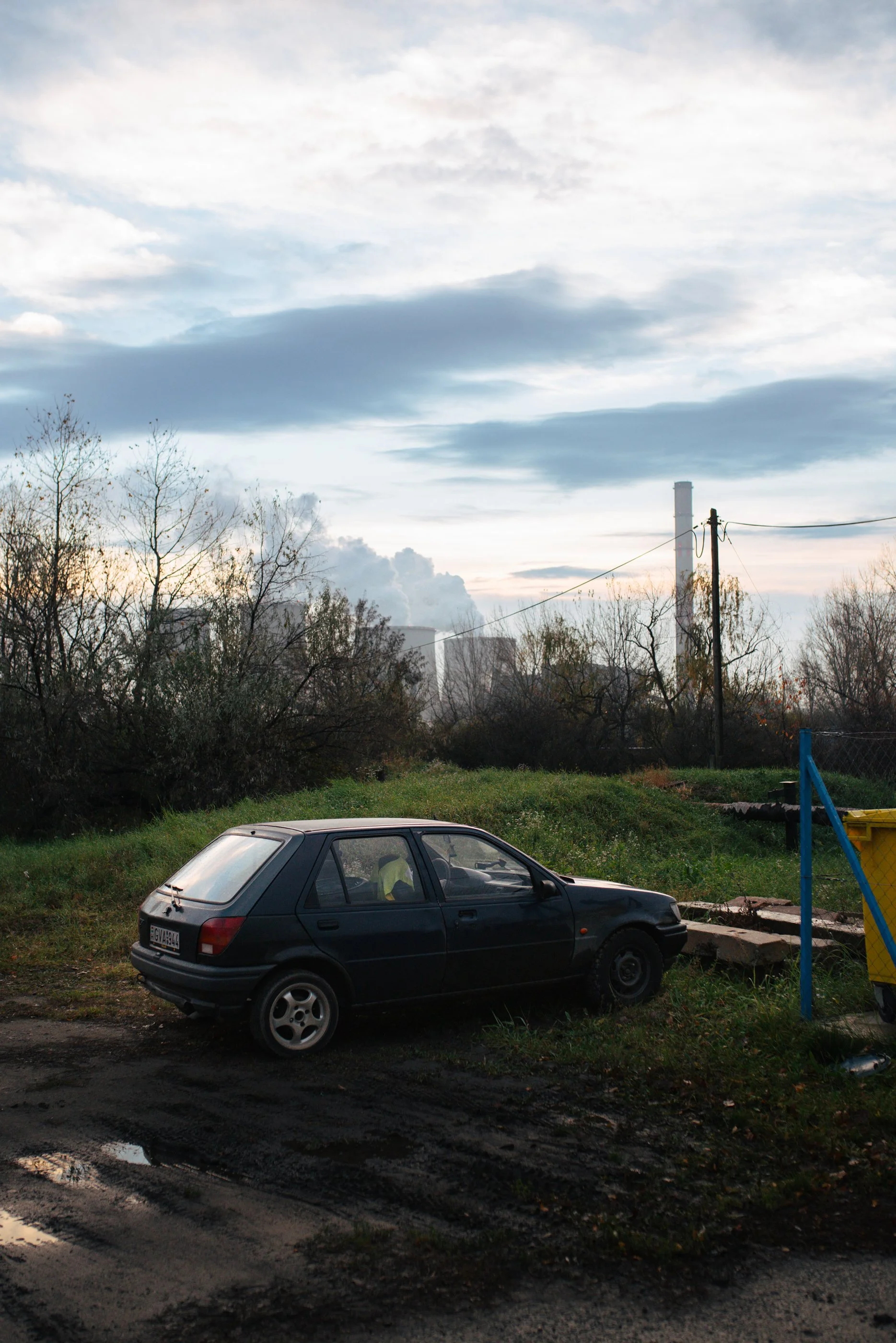 Ein schwarzes Auto steht auf einer unbefestigten Straße neben einer Wiese mit Buschwerk und Bäumen, mit einem Industriegebiet im Hintergrund unter einem bewölkten Himmel.