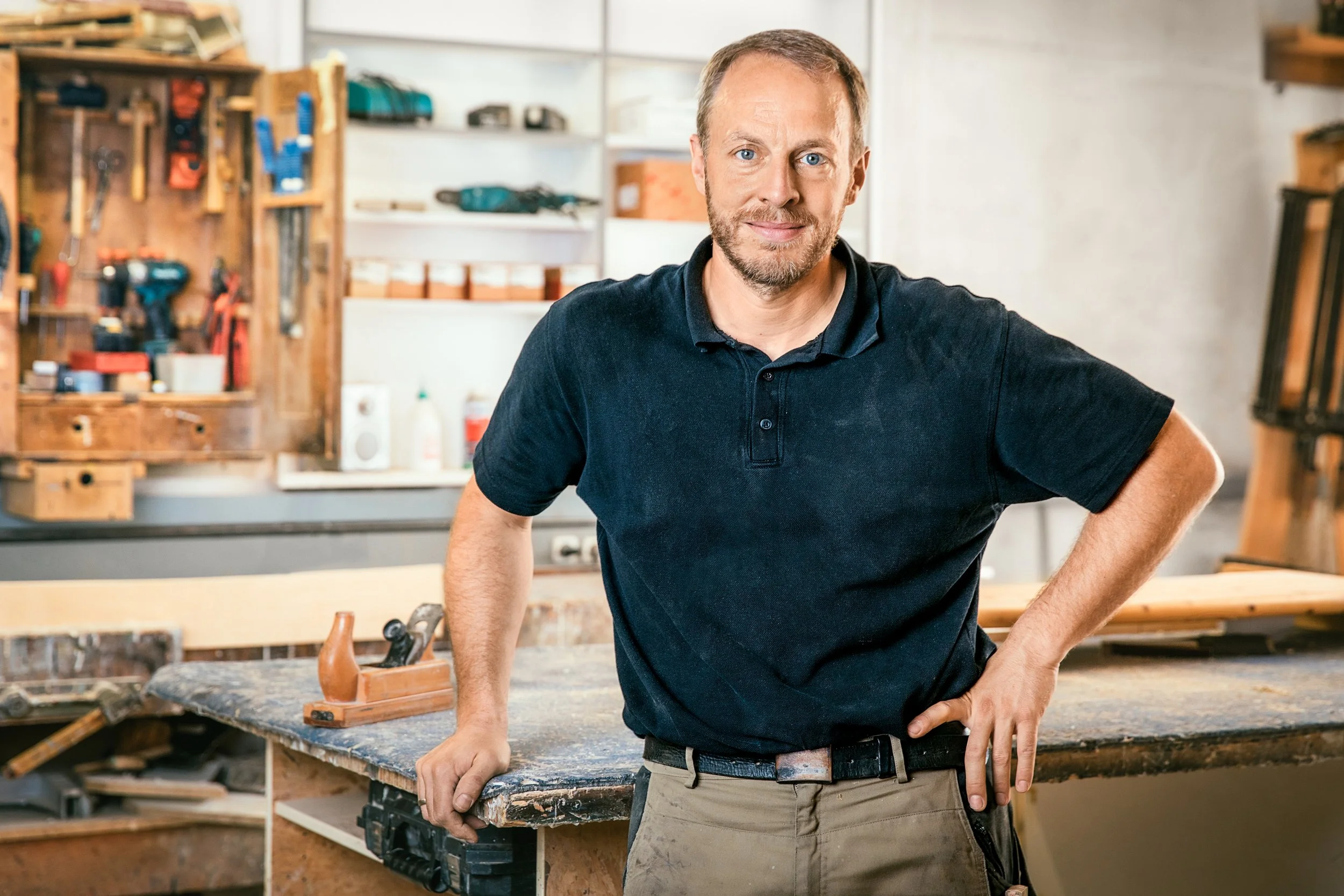 Ein Mann in einer Multimediwerkstatt steht vor einer Werkbank mit Werkzeugen, lächelt und trägt ein schwarzes Polo-Shirt und beige Arbeitshosen.