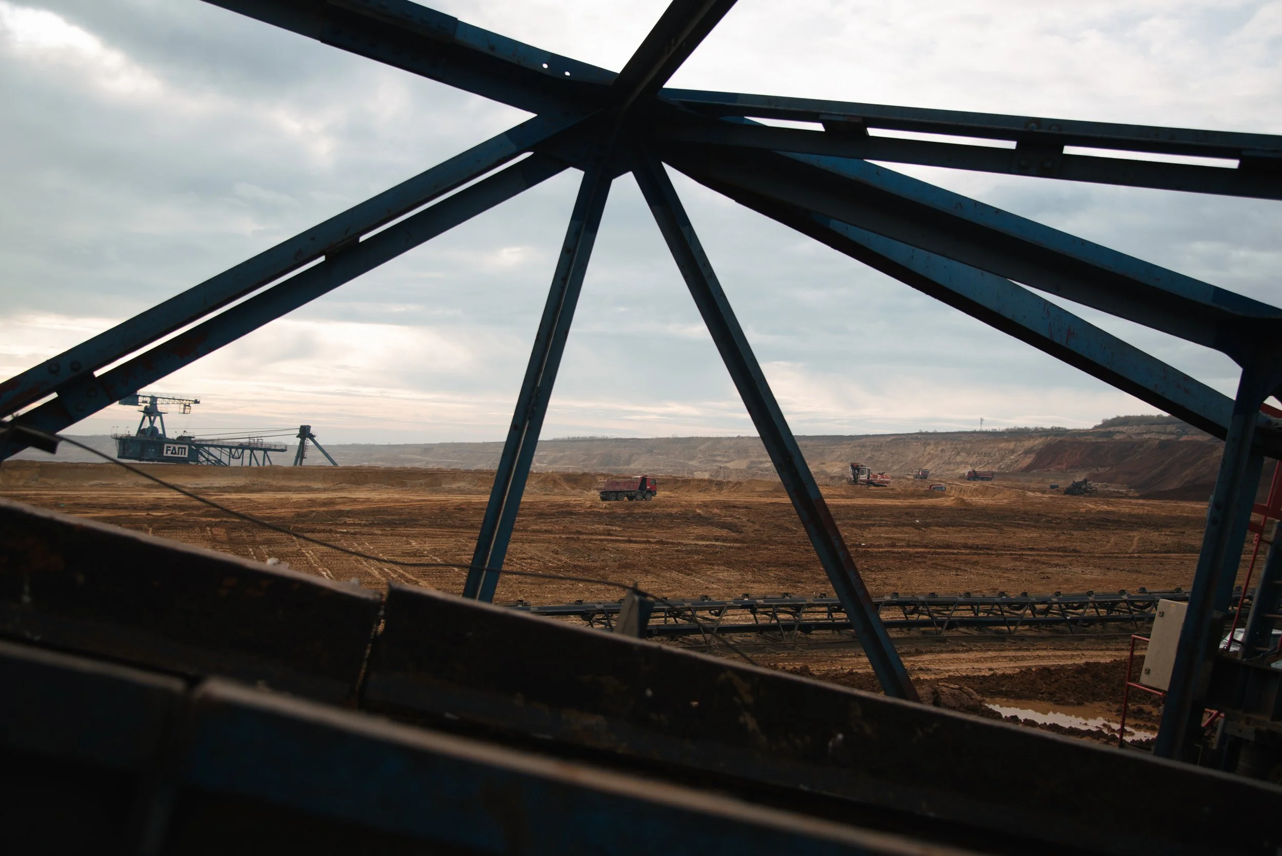 Blick auf eine riesige Industrie- oder Bergbaulandschaft mit schwerem Gerät und Dampfwolken am Horizont, sichtbar durch eine Metallstruktur im Vordergrund.