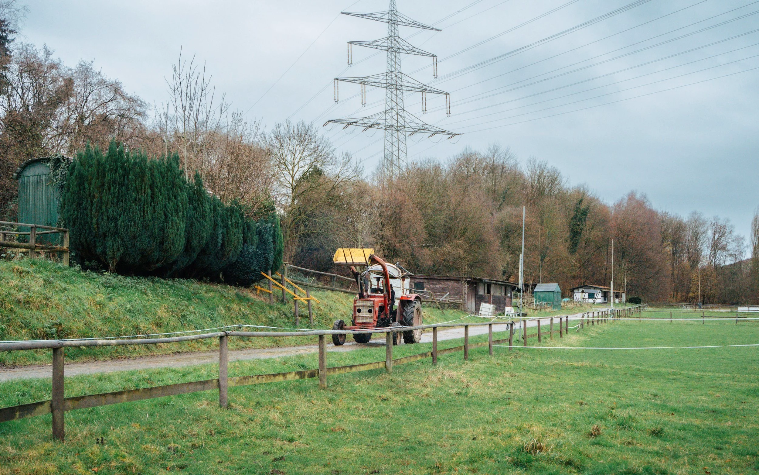 Blick auf eine ländliche Szene mit einem Traktor auf einem Weg, umgeben von Grünflächen, Büschen und Bäumen, mit Strommasten im Hintergrund.