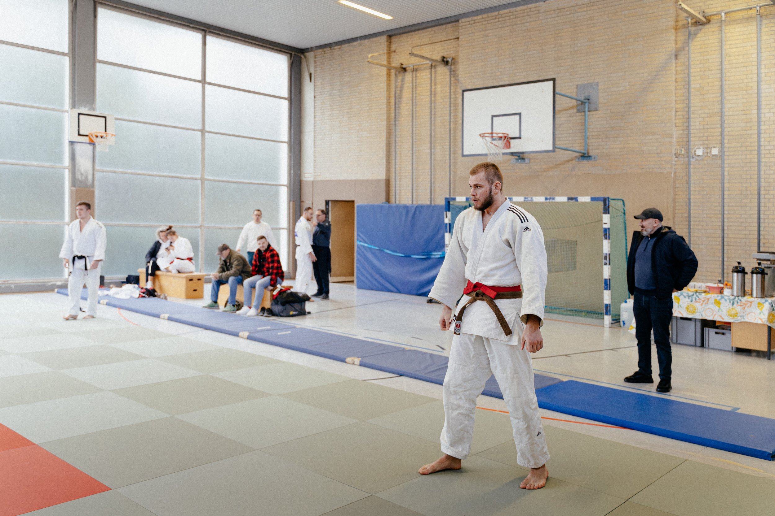 Junge Männer in Judogis beim Judo-Training in einer Turnhalle, einige sitzen auf Bänken, andere stehen, im Hintergrund sind Sportgeräte und Zuschauer.