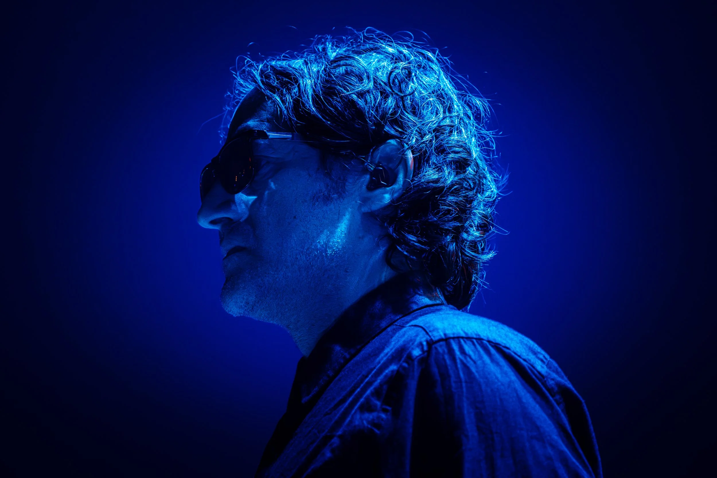 Ein älterer Mann mit grauem, lockigem Haar trägt schwarze Sonnenbrille und ein dunkles Hemd, präsentiert in blauer Beleuchtung in einem künstlerischen Stil.