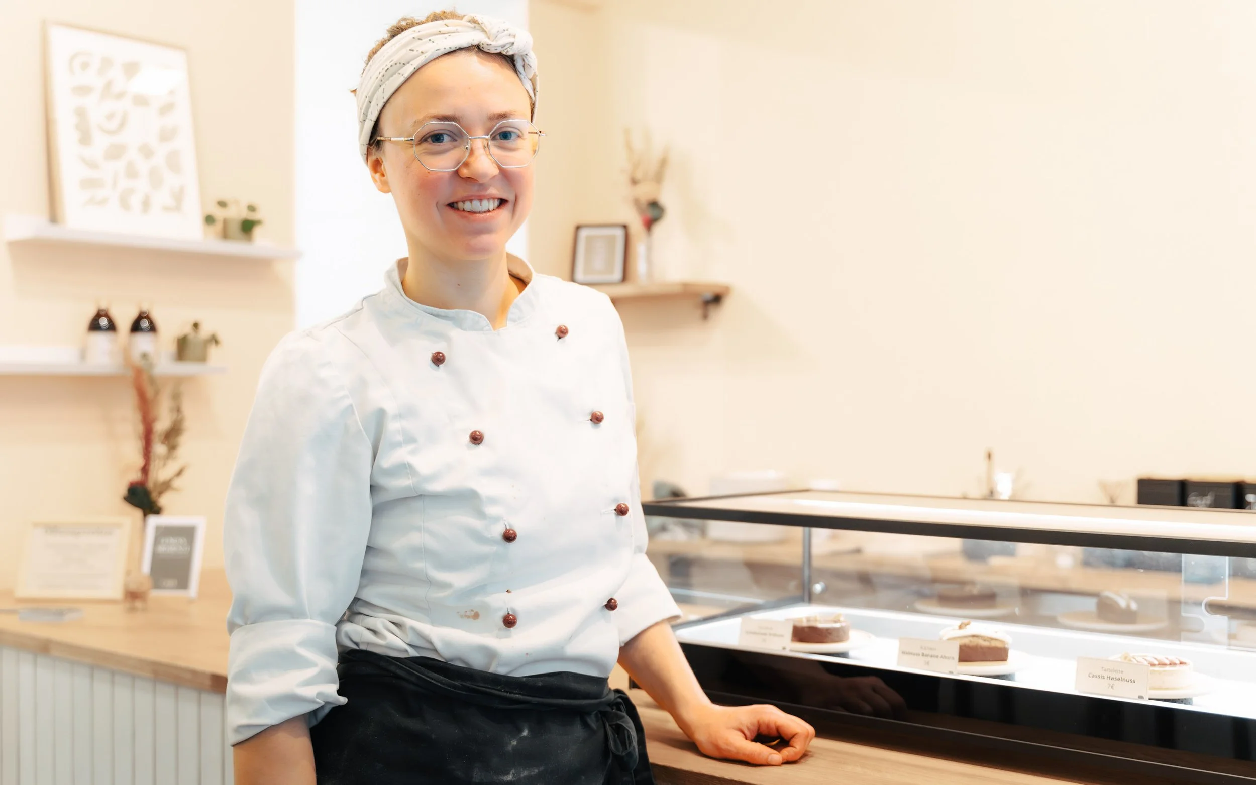 Küchenchefin in einem Café oder einer Patisserie, die hinter einer Glasvitrine mit Kuchen steht, trägt weiße Kochjacke, Kopfband, Brille und lächelt. Im Hintergrund sind Dekorationsgegenstände an der Wand.