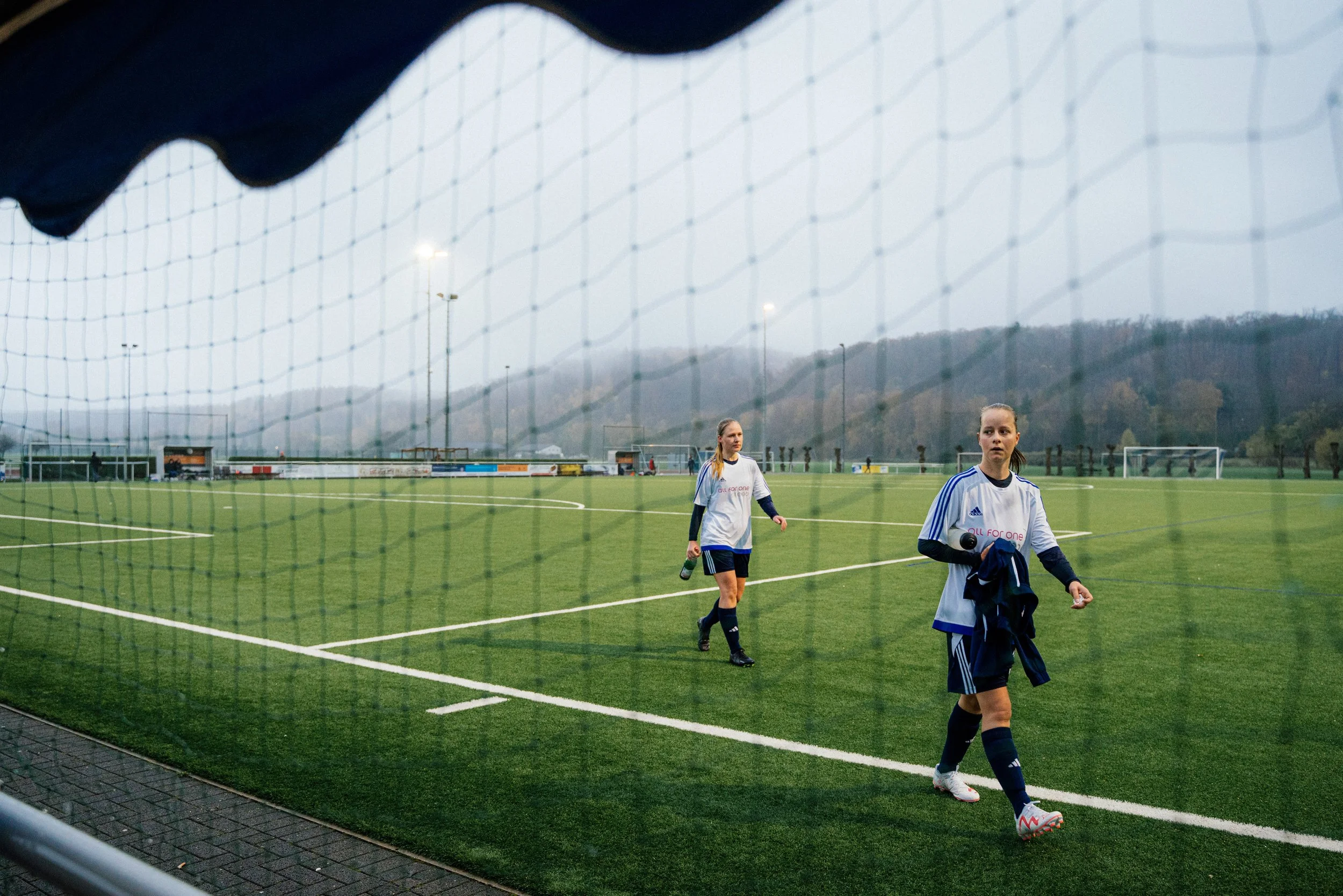 Zwei weibliche Fußballspielerinnen auf einem grünen Sportplatz, eine hält eine Wasserflasche, die andere trägt Kleidung. Im Hintergrund sind Torpfosten und Hügel zu sehen, der Himmel ist bewölkt.