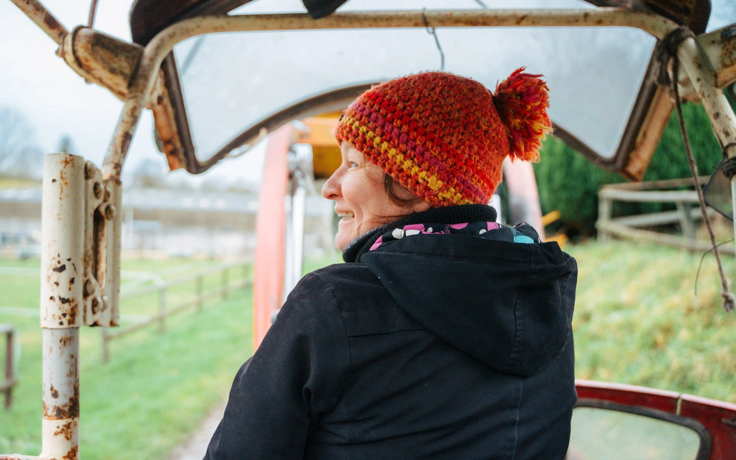 Frau mit roter, gestrickter Mütze, lachend, im offenen Korb eines landwirtschaftlichen Fahrzeuges, im Hintergrund grünes Feld und Bäume, bedeckter Himmel.