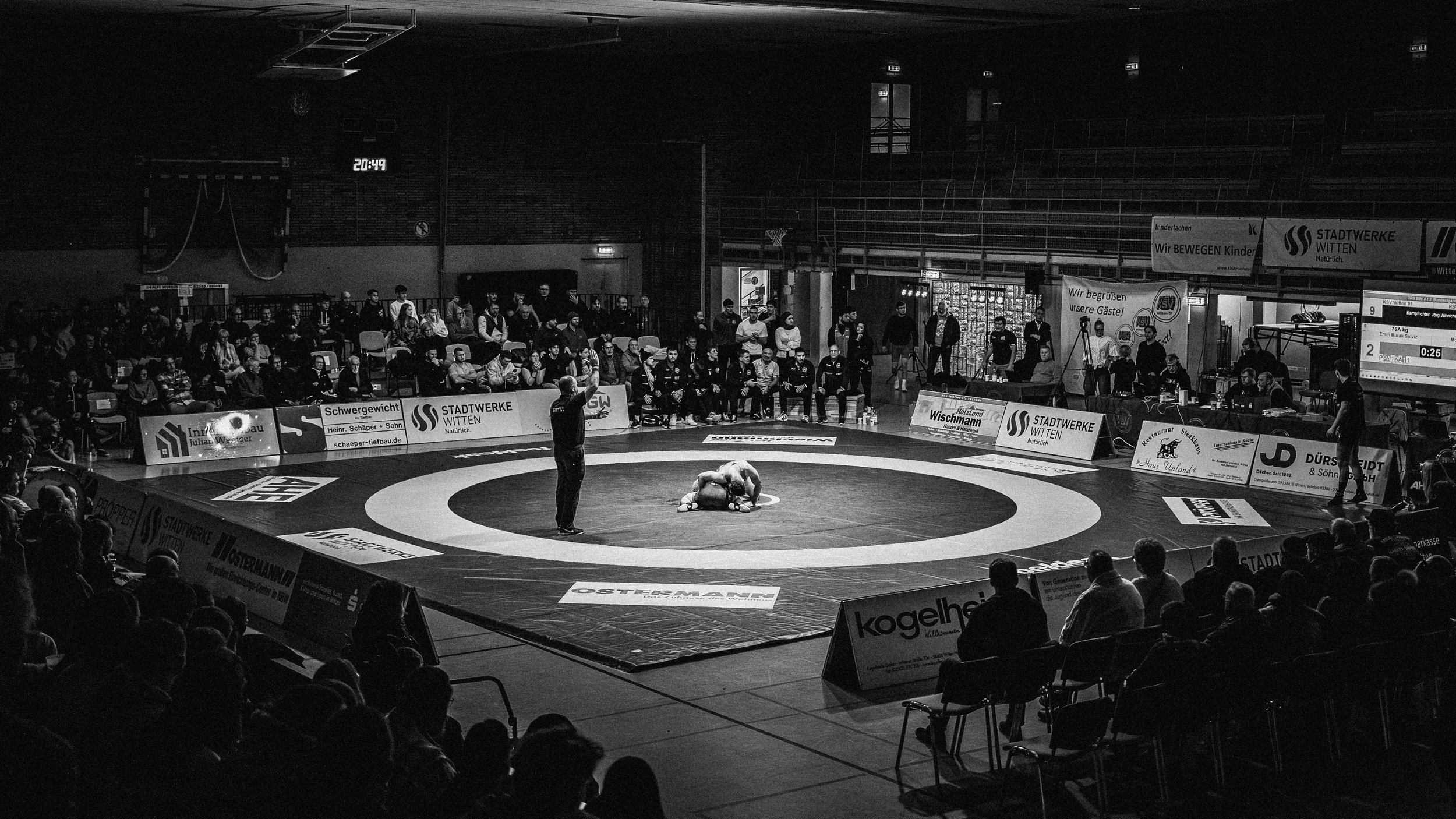 Zwei Ringer im Wrestling auf einem Turnier in einem Turnring, umgeben von Zuschauern in einer Halle, Schwarz-Weiß-Fotografie.