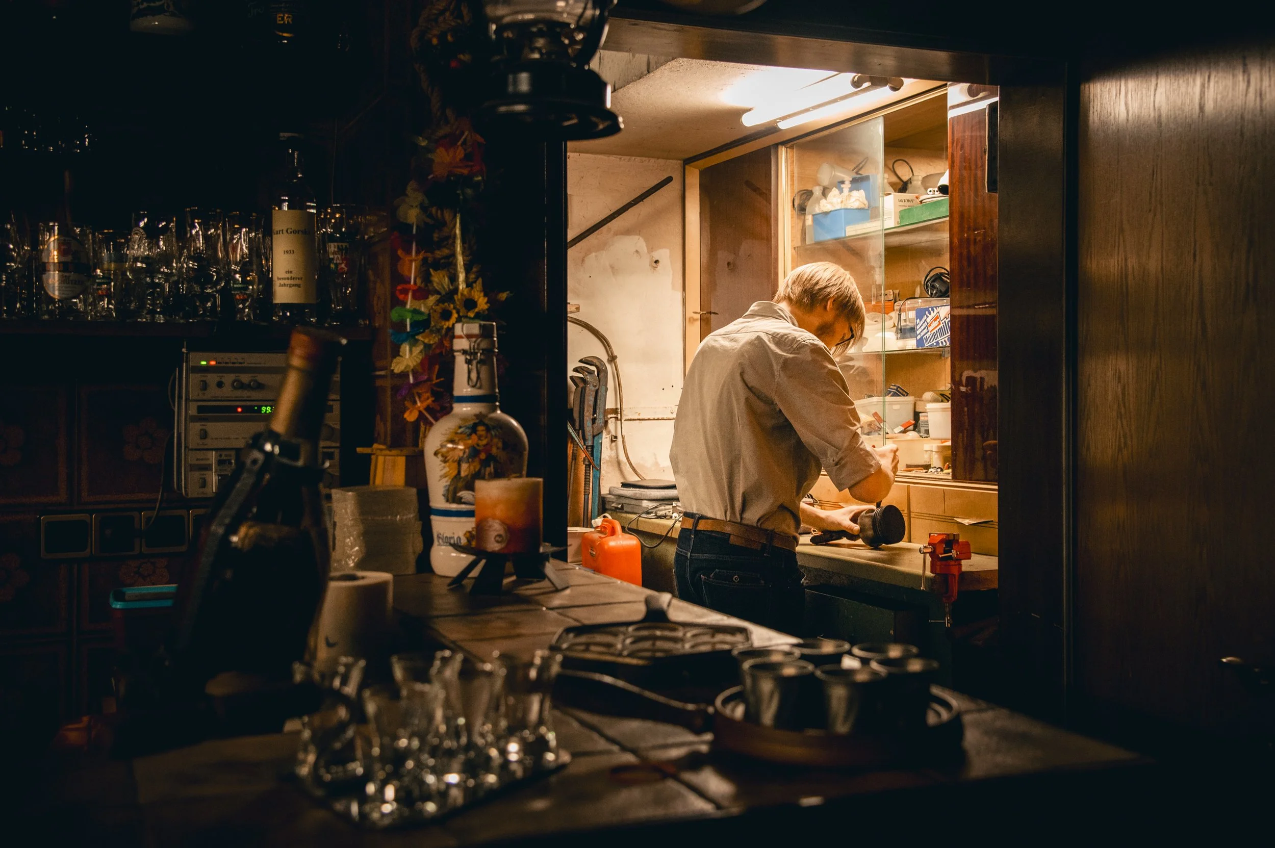 Ein junger Mann arbeitet in einer kleinen Küche oder Bar bei schwachem Licht, umgeben von Flaschen und Küchenutensilien.