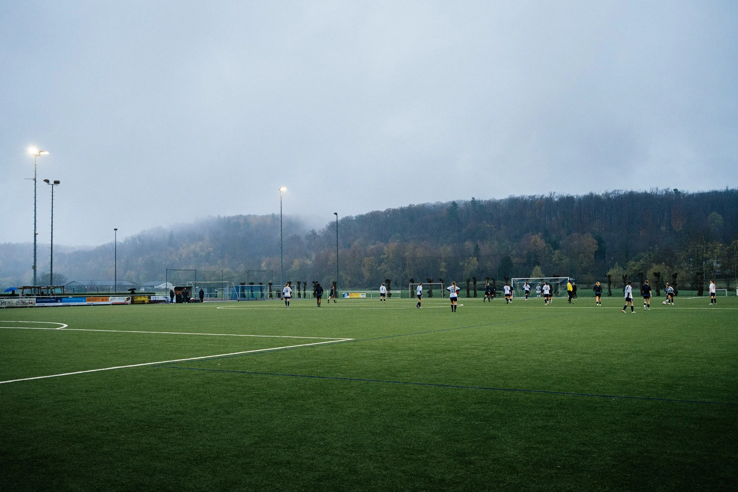 Fußballspiel auf einem Rasenplatz bei bewölktem Himmel, mit Spielern in weißen und dunklen Trikots, im Hintergrund Wald und Bergland, bei Nacht die Flutlichtmasten beleuchtet.
