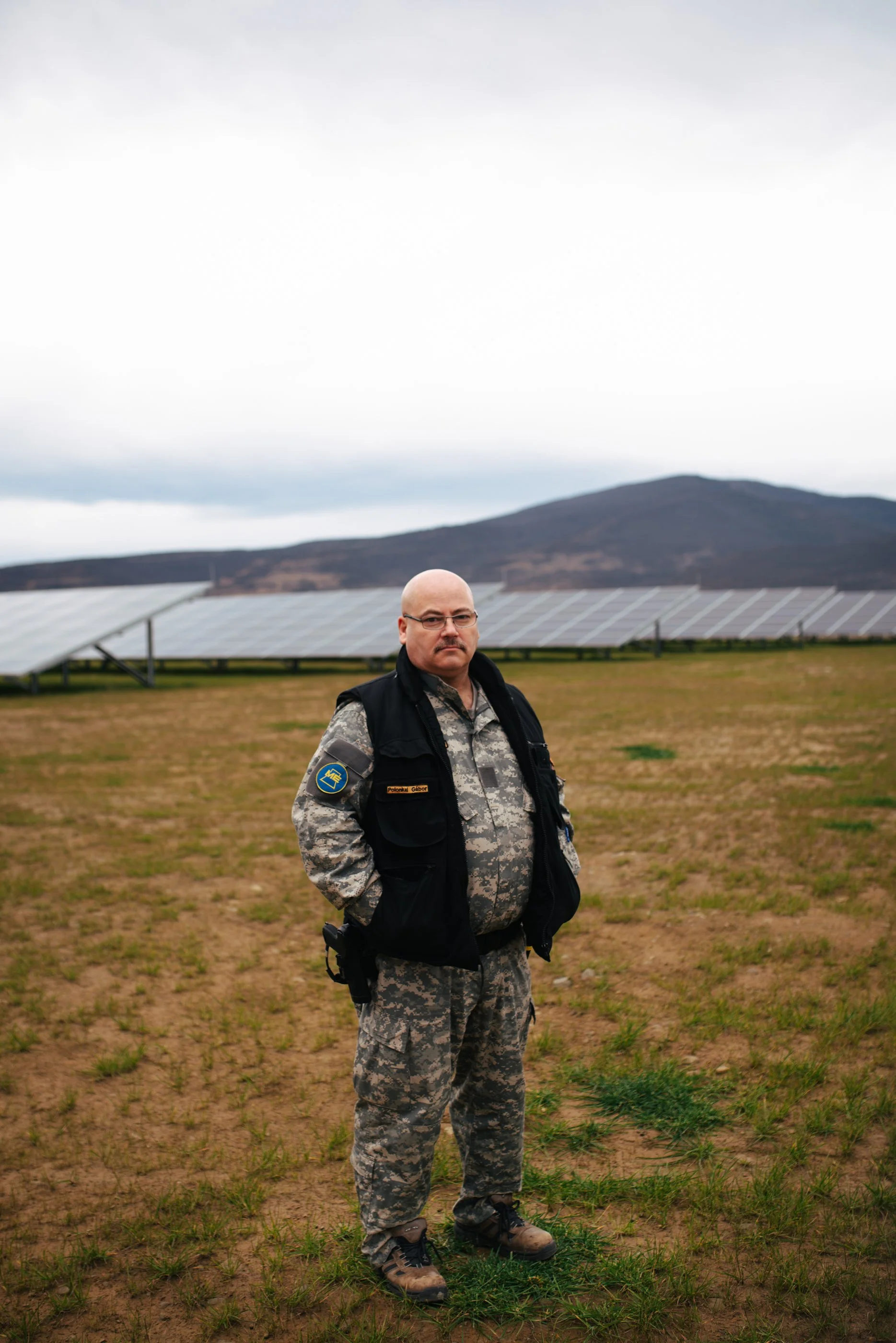 Ein Mann in Camouflage-Uniform mit schwarzen Weste, Sonnenbrille und sanftem Blick steht auf einem Feld vor einer Solaranlage mit Bergen im Hintergrund.