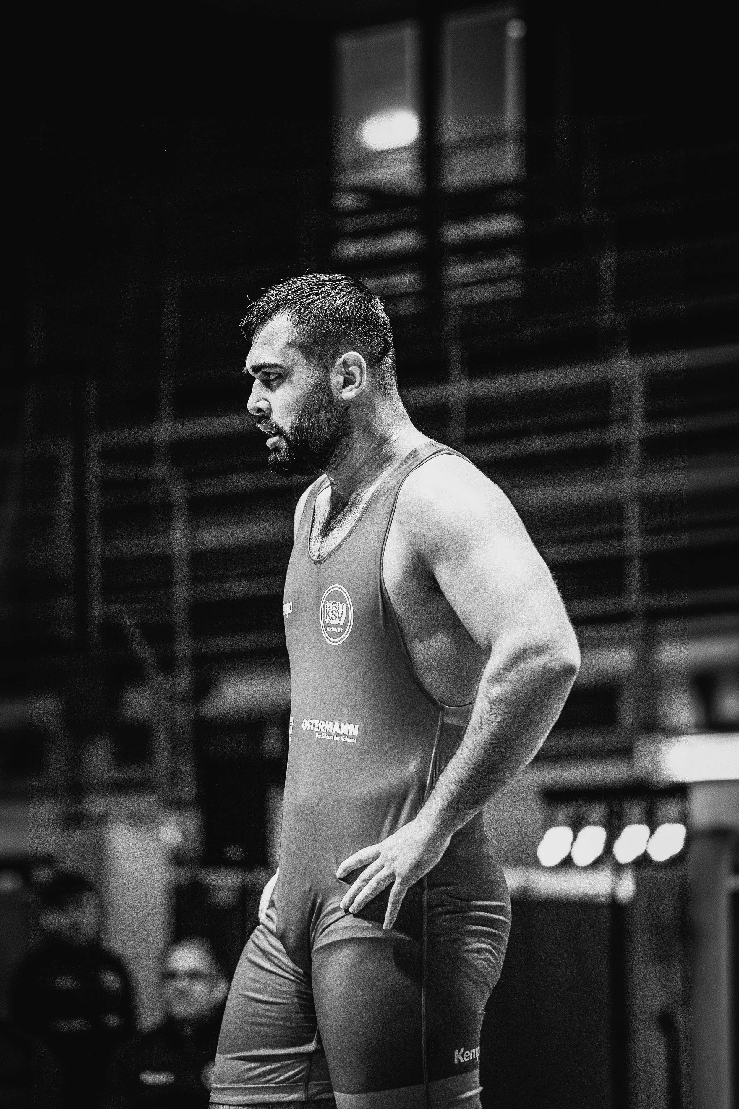 Schwarz-weiß-Foto eines männlichen Athleten in einem Sporthaus, trägt einen Trainingsanzug, wirkt konzentriert und nachdenklich.