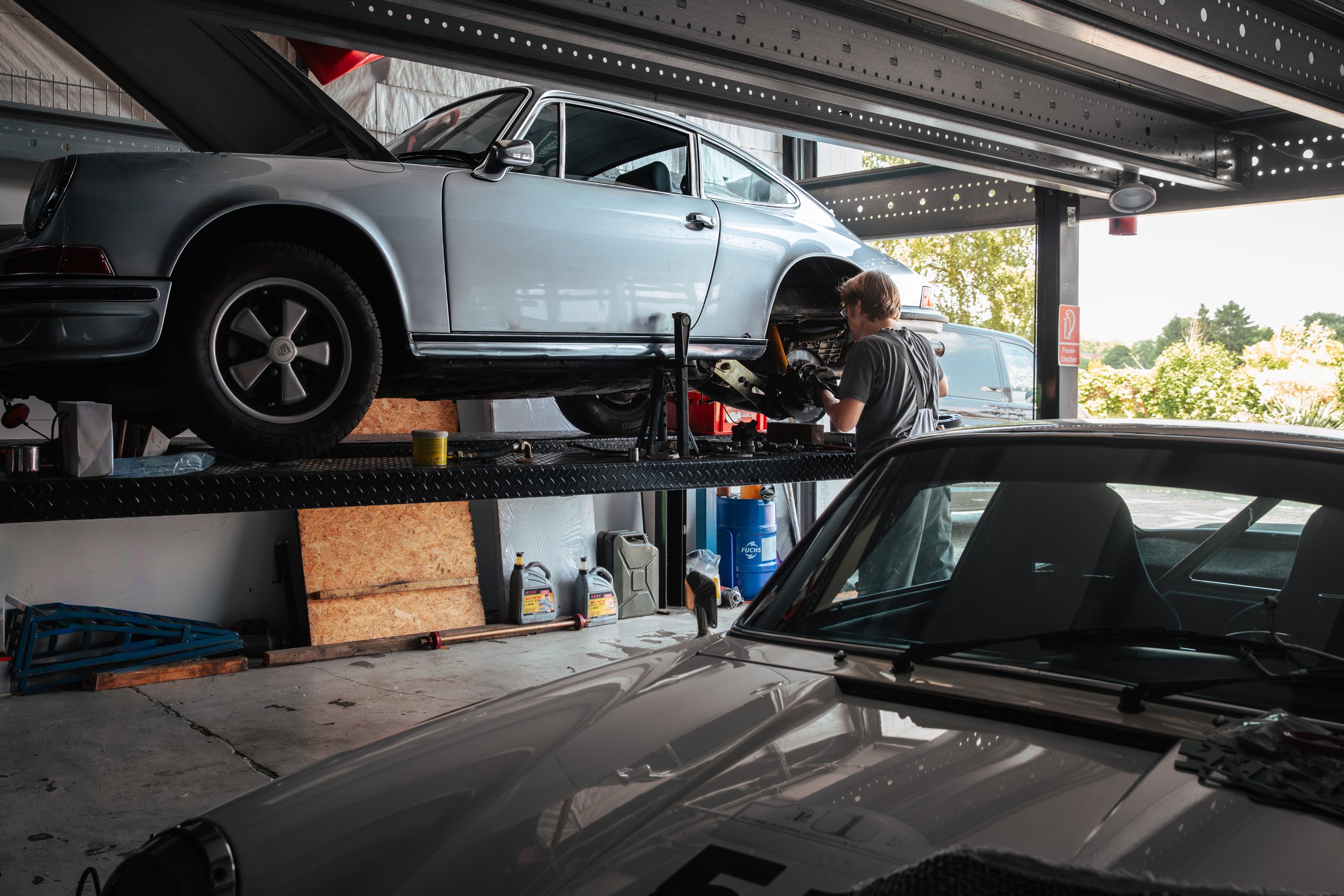 Ein Mechaniker arbeitet an einem halbhoch stehenden weißen Porsche im Autowerkstatt, während im Vordergrund ein silberner Oldtimer zu sehen ist.
