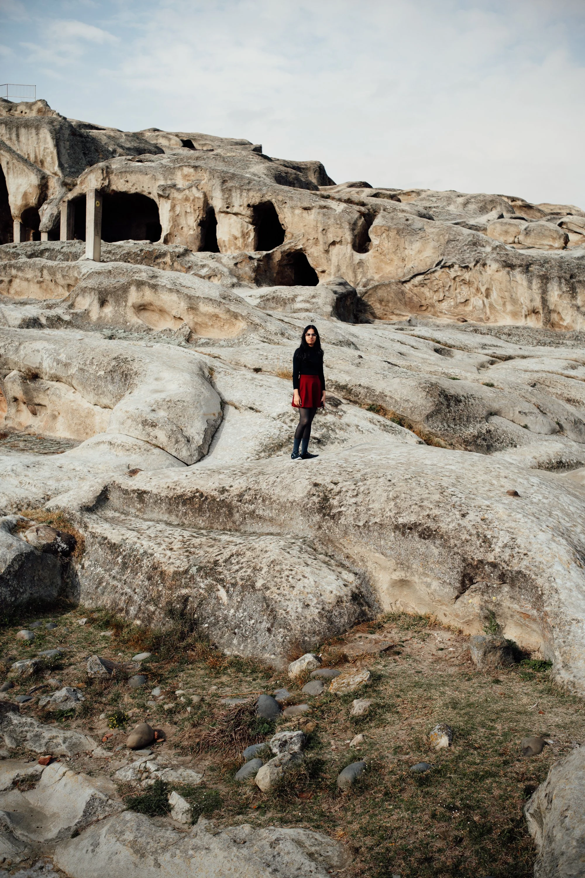 Eine Frau steht auf einem felsigen Gelände vor einer ausgetrockneten, steinigen Landschaft mit Höhlen und Felsformationen im Hintergrund.