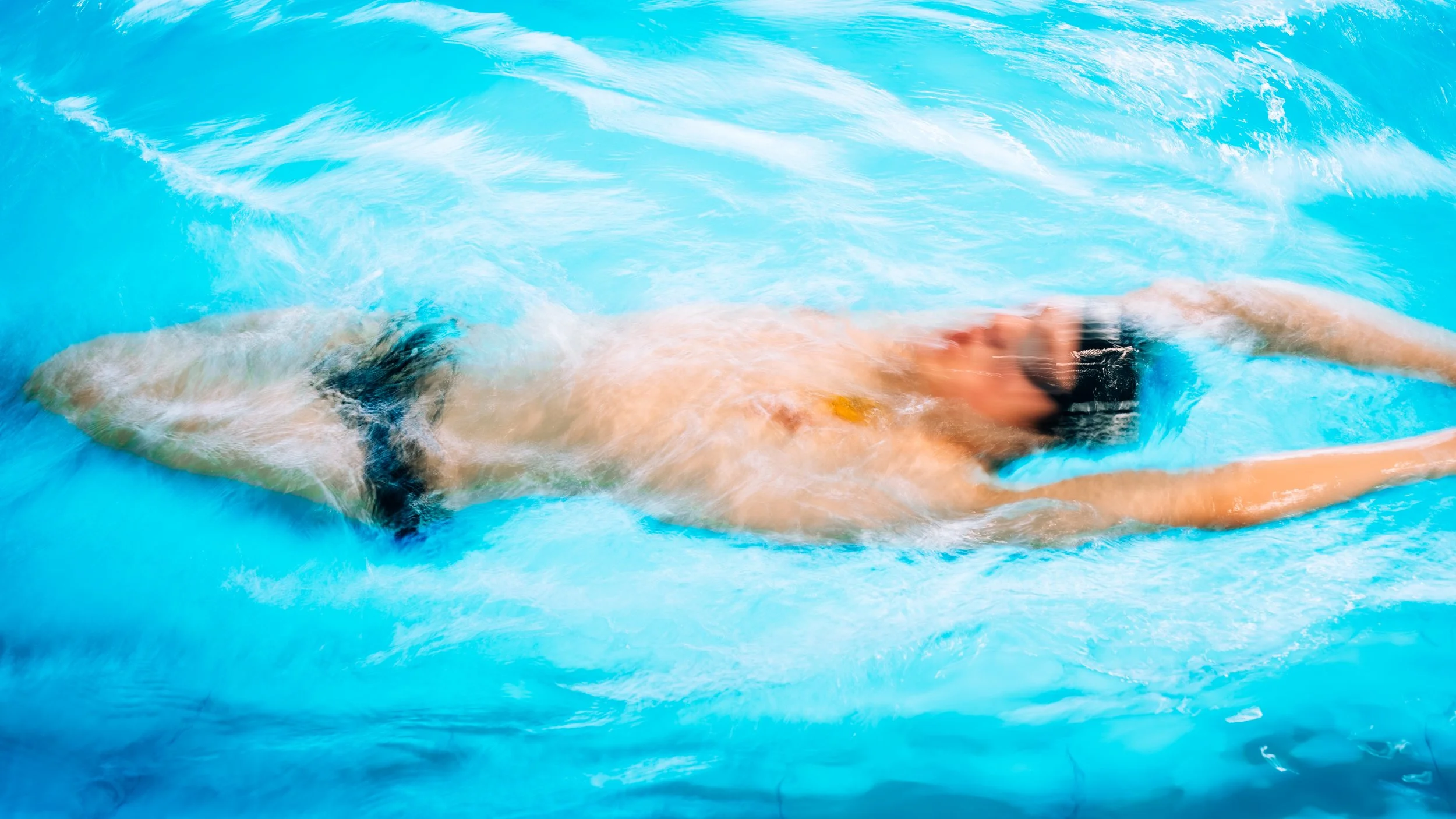 Schwimmer im Wasser beim Wettkampf, im Wasser liegend, arm nach vorne streckend, im Pool mit blauer Wasserfläche.