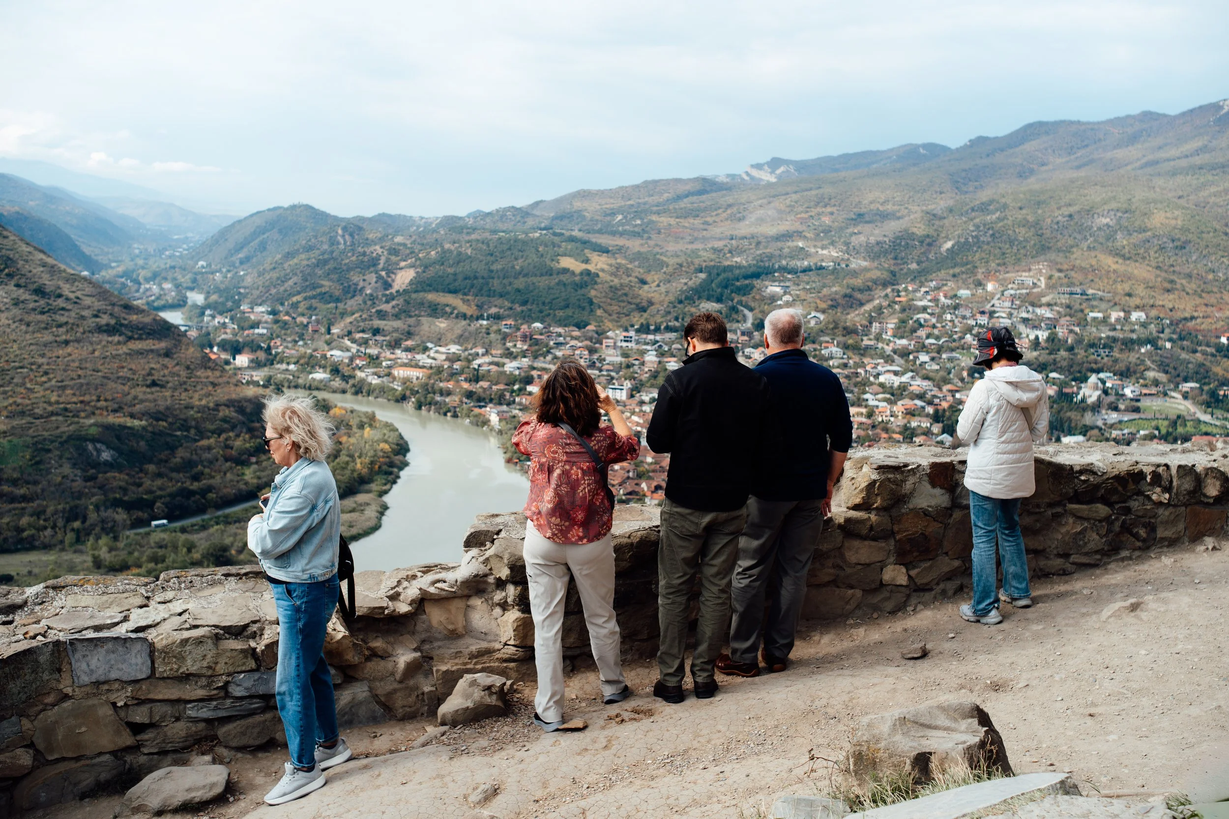 Menschen stehen an einer Steinmauer und blicken auf eine Flusslandschaft mit Stadt, bewaldeten Hügeln und Bergen in der Ferne.