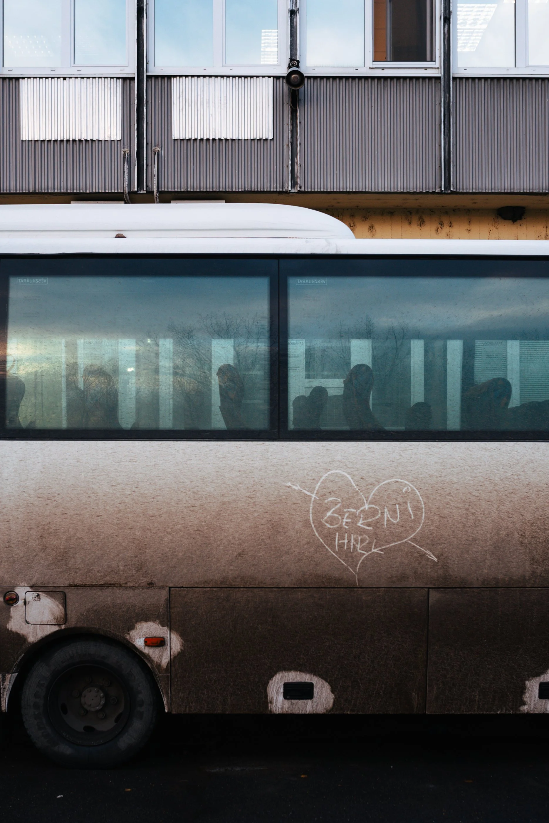 Ein verchromter Bus mit reflektierenden Fenstern, auf dem eine Herz-Schablone mit der Aufschrift "BEHÜT HIR" in Kreide gemalt ist, parked vor einem modernen Wohngebäude.