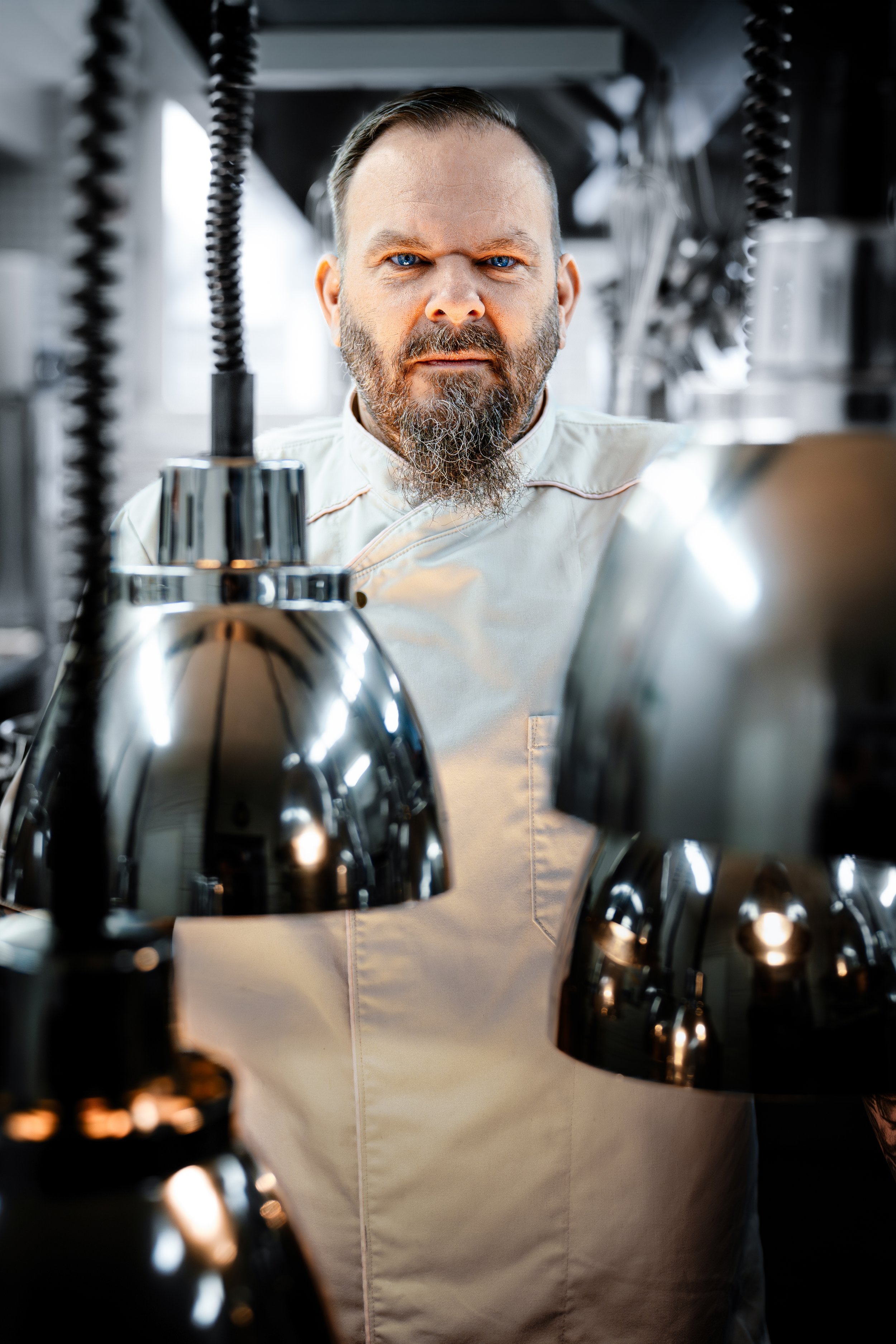 Koch in einer Küche, schaut ernsthaft in die Kamera, umgeben von glänzenden, metallischen Küchenutensilien.