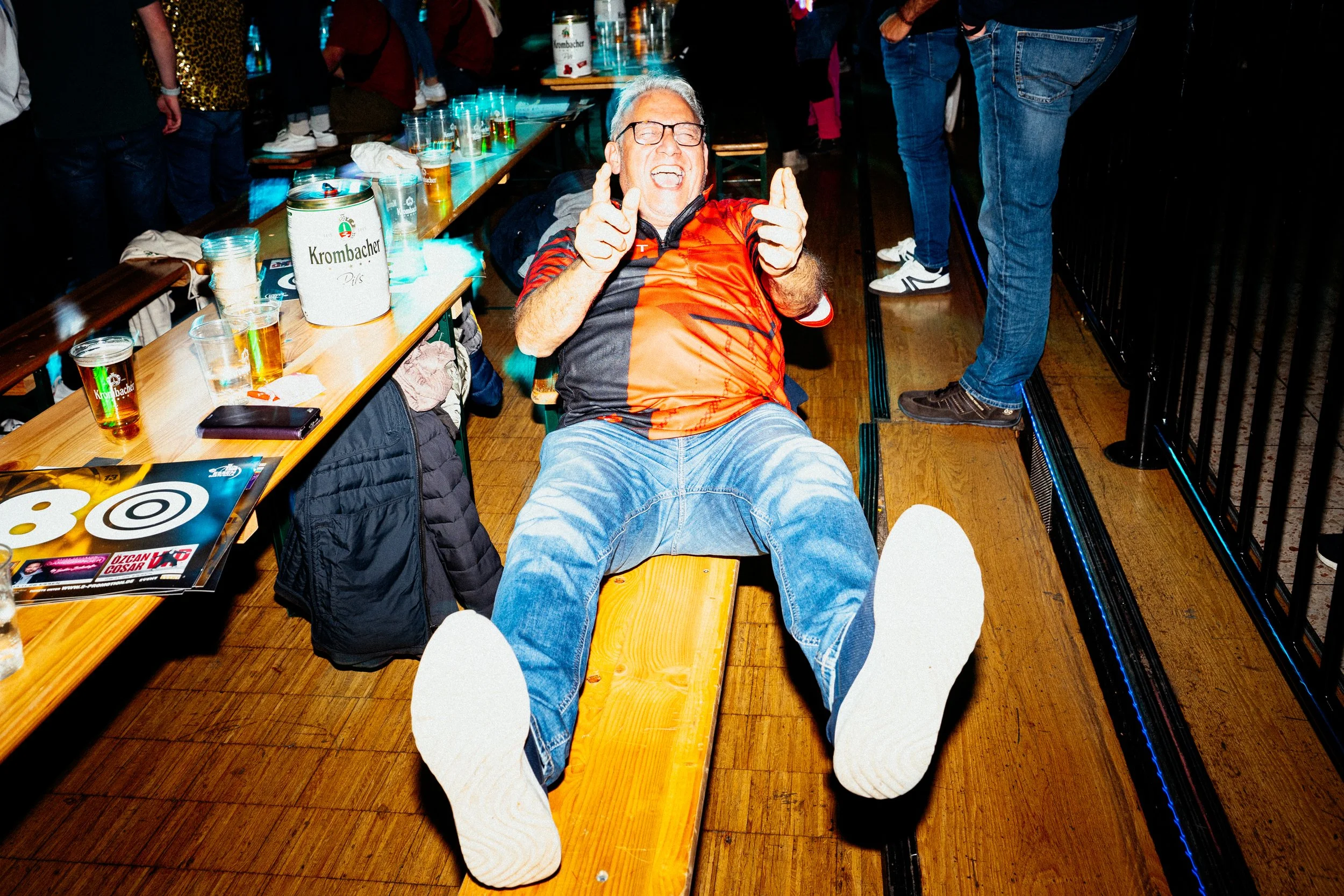 Ein lachender Mann sitzt auf einem Stuhl, dessen Beine auf einem Holz-Klapptisch liegen, in einer Bar oder beim Event, umgeben von leeren Biergläsern und Getränkekästen, während er in die Kamera zeigt.