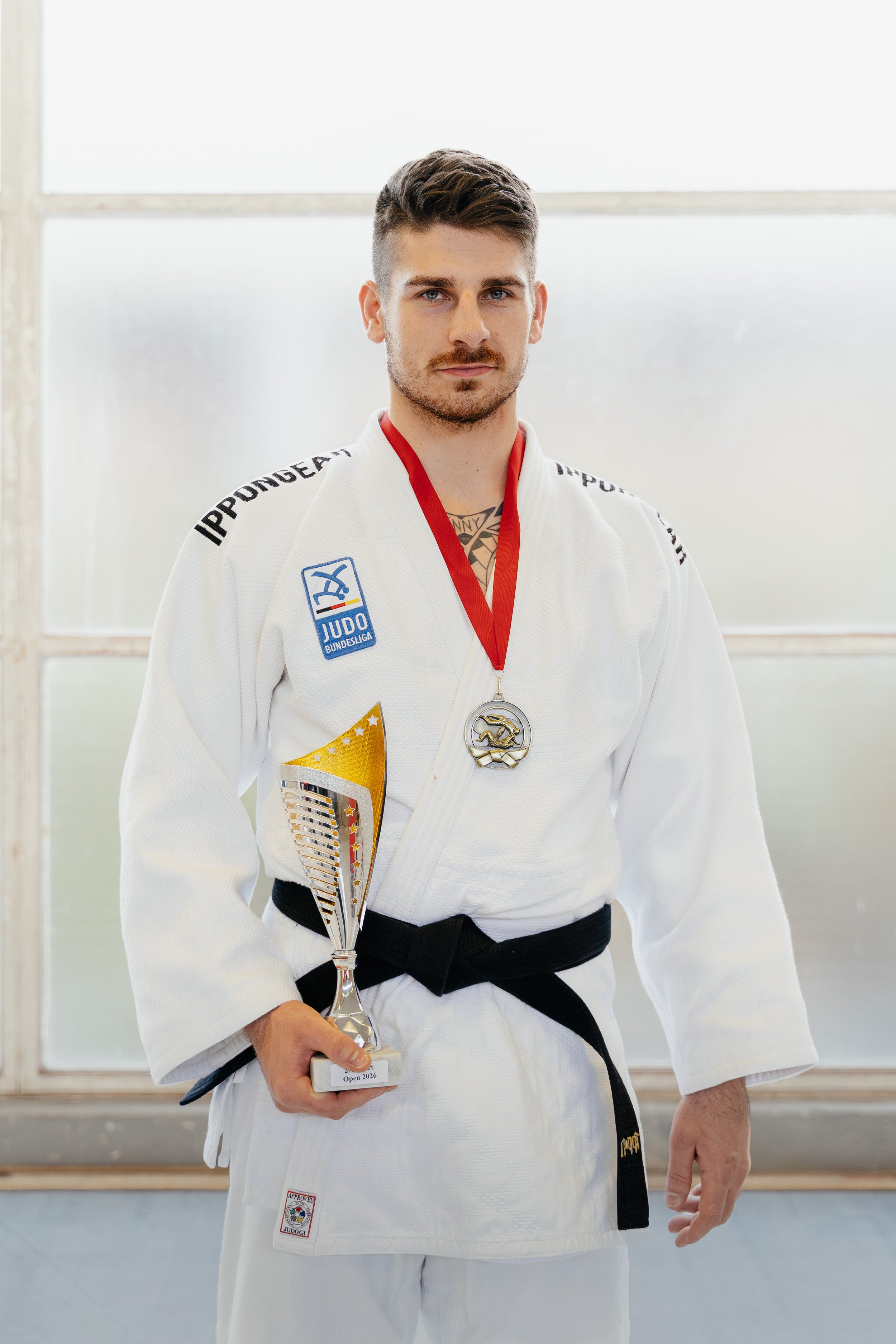 Ein junger Mann in Judo-Gi mit schwarzen Gürtel hält einen Pokal und trägt eine Medaille um den Hals, vor einem Fenster