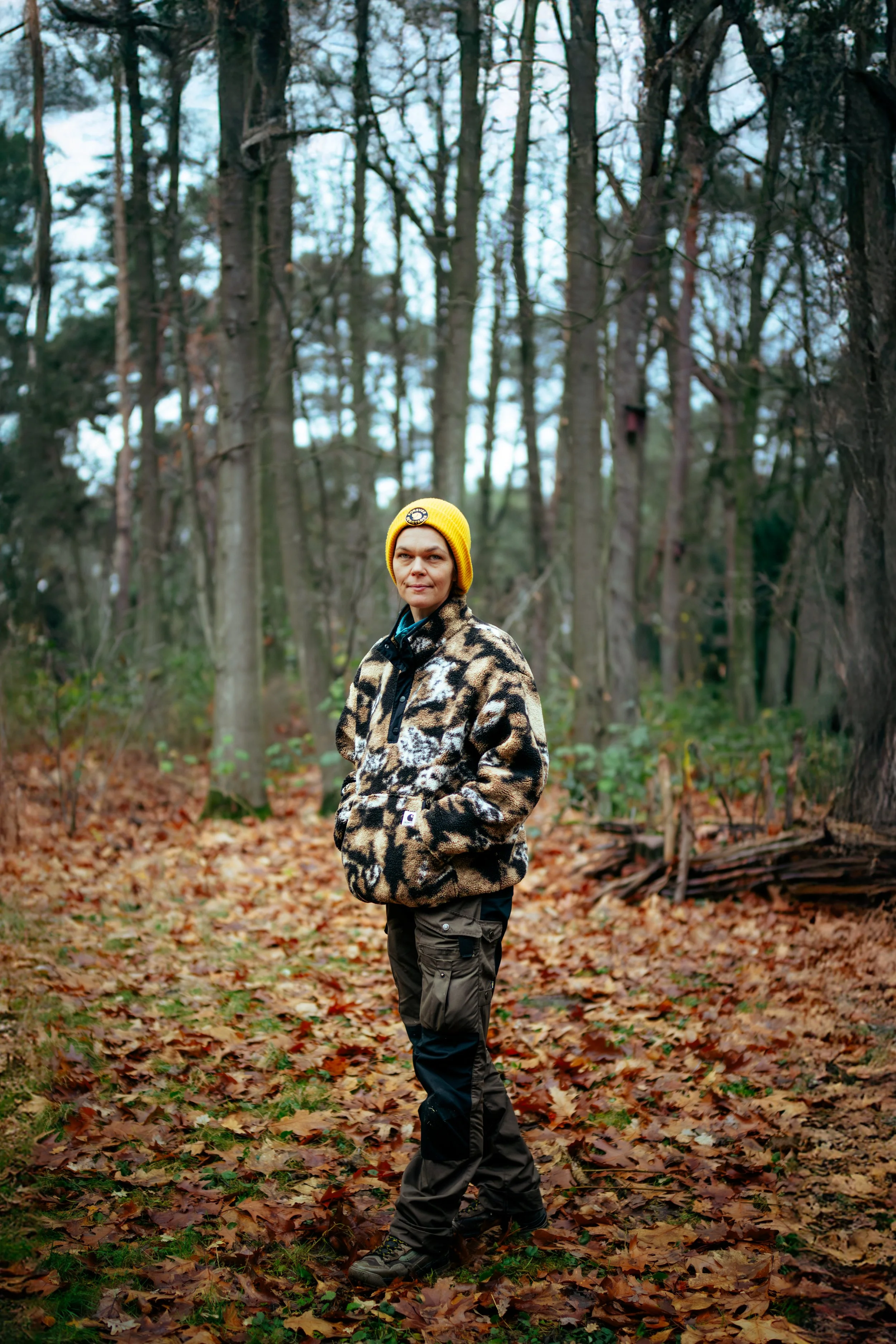 Frau in Park mit Laub auf dem Boden, trägt karierte Jacke und gelben Hut, steht im Wald.