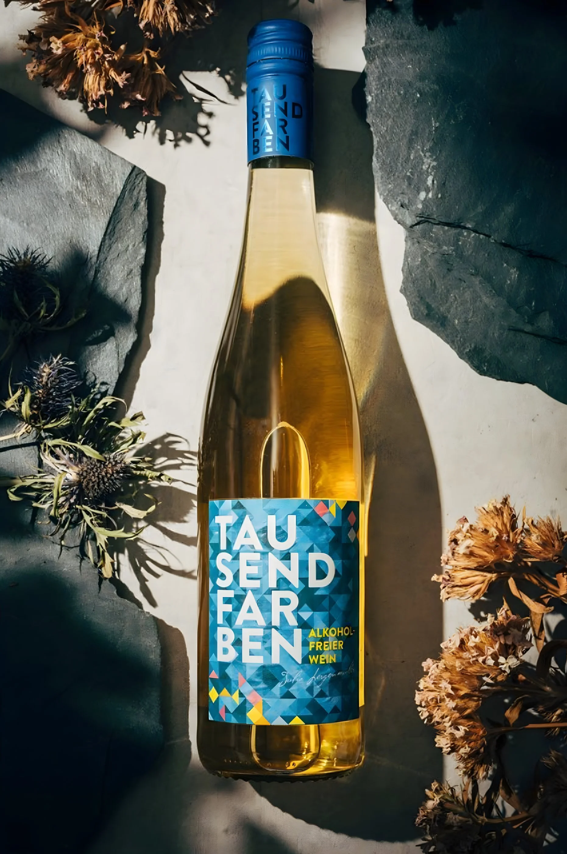 Bunte Flasche mit der Aufschrift "Tau Send Farben" auf einem Tisch, umgeben von getrockneten Blumen und Steinen.
