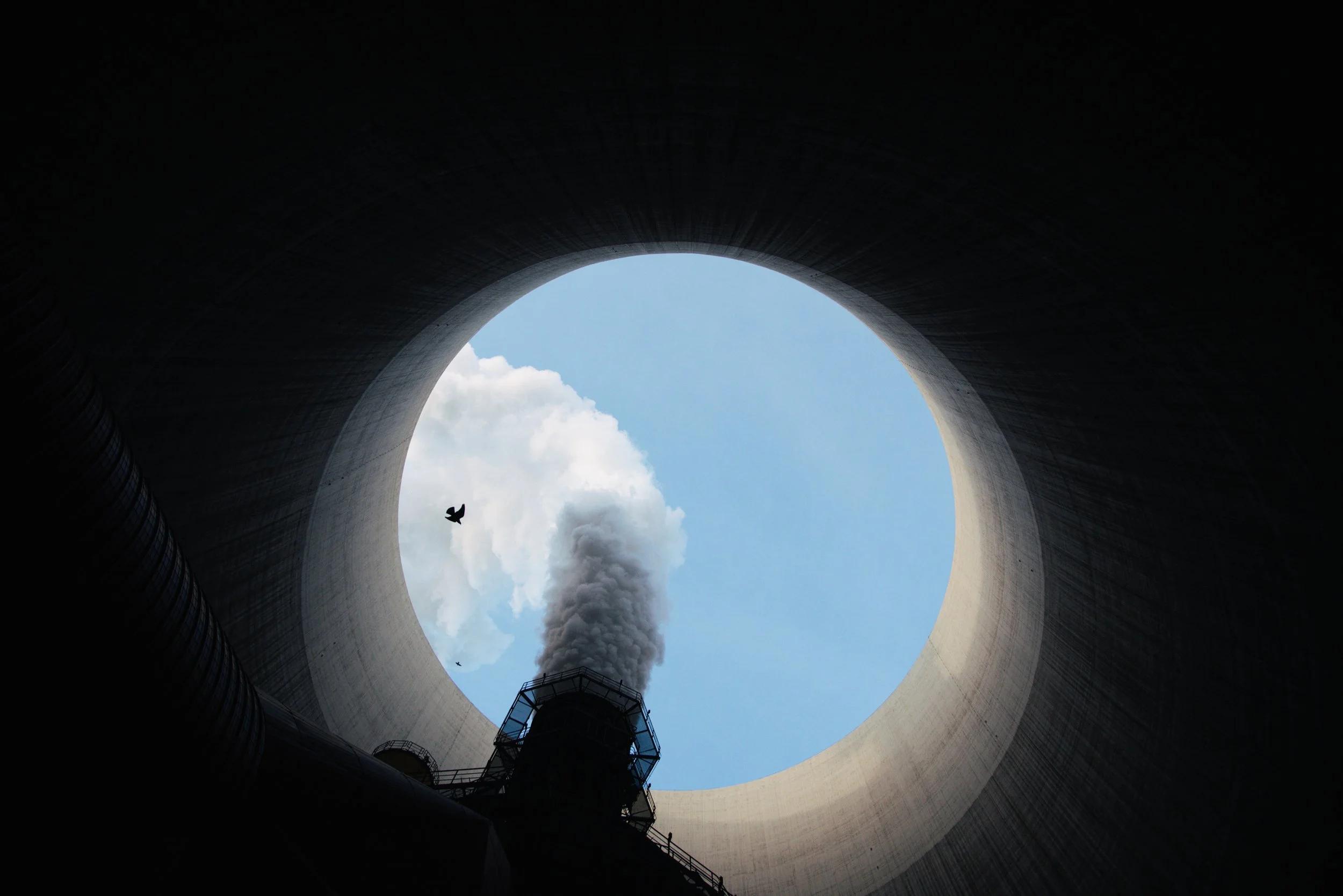 Blick nach oben in ein industrielles Kühlturmloch, das den Himmel und Wolken zeigt, mit aufsteigendem Rauch und einem Vogel.