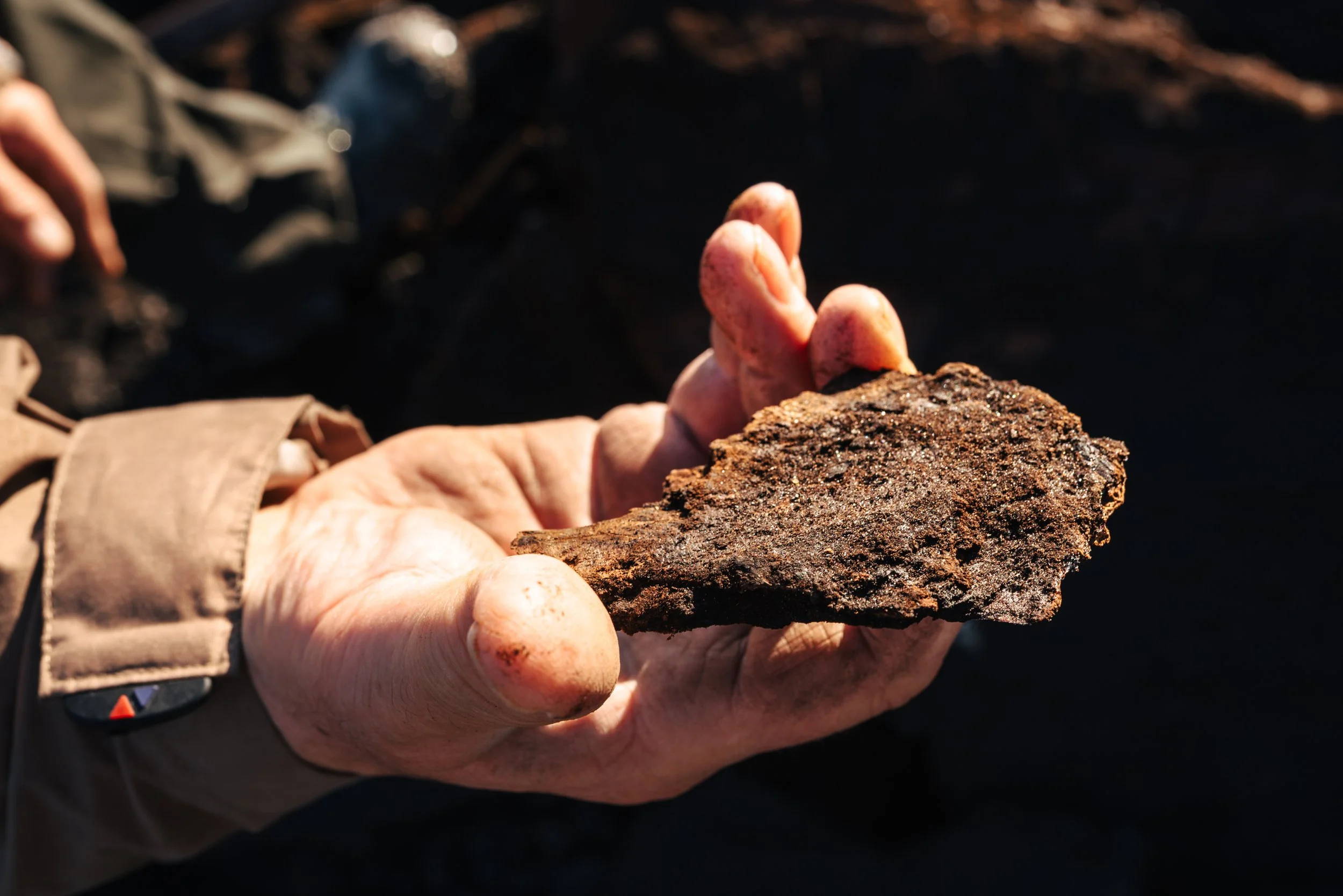 Eine Hand hält einen rostigen, alten Metallgegenstand, möglicherweise ein Werkzeug, vor dunklem Hintergrund.