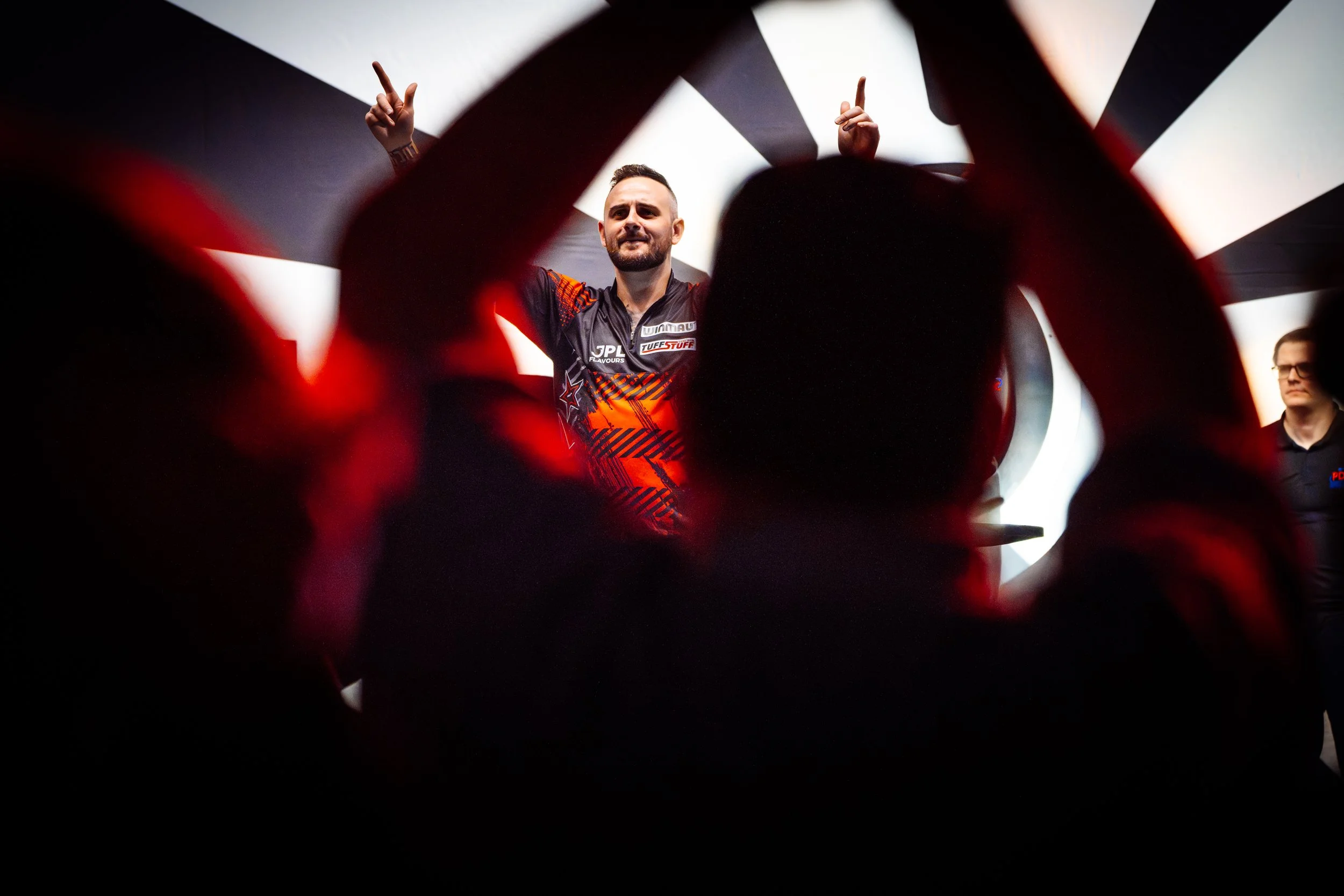 Ein Mann in einem schwarzen und orangefarbenen Sporttrikot steht auf einer Bühne, umgeben von Silhouetten von Menschen mit erhobenen Armen, die das Bild umrahmen.