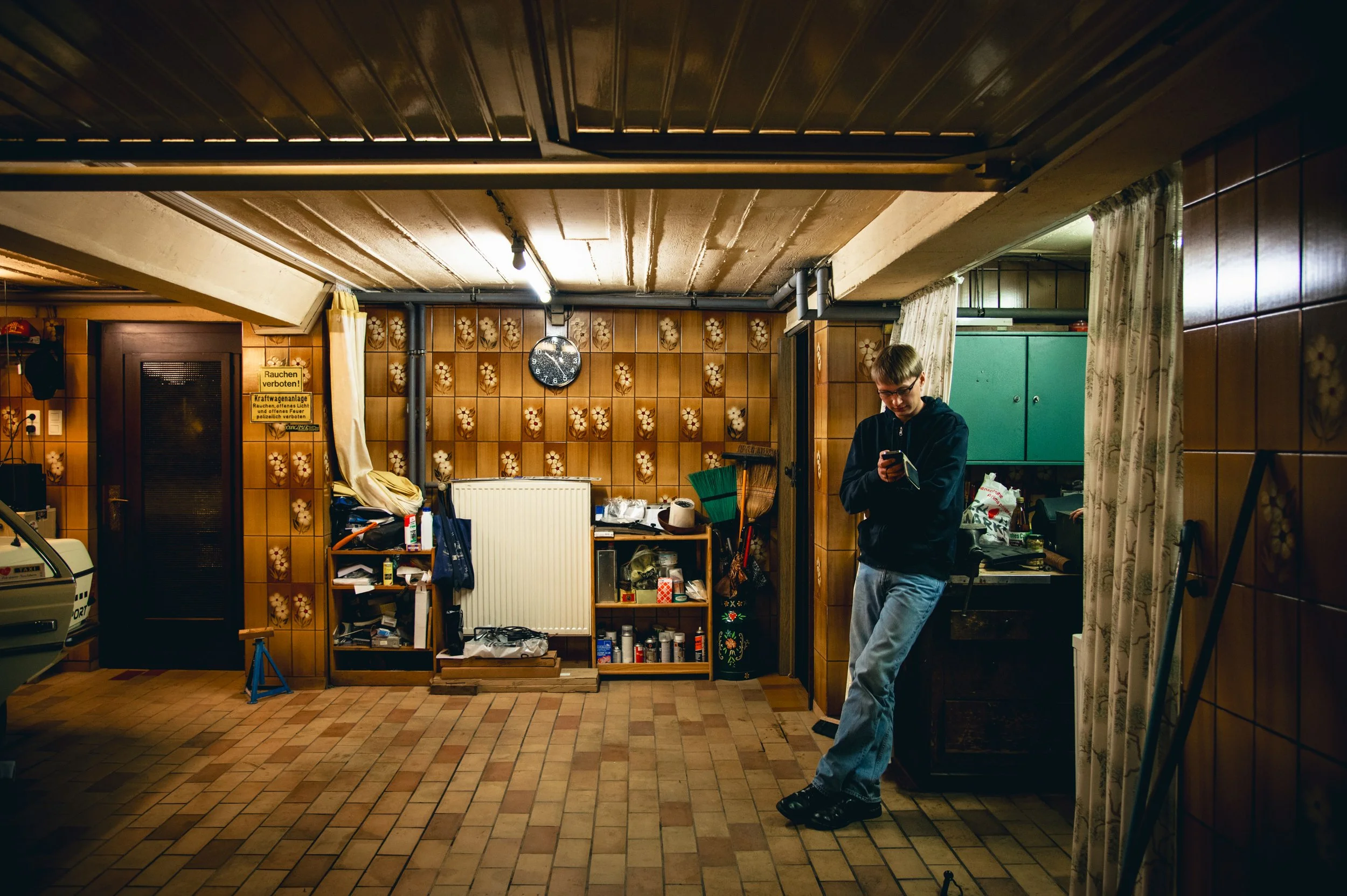 Junge Person lehnt an einer Wand in einem Kellerraum und schaut auf sein Smartphone.