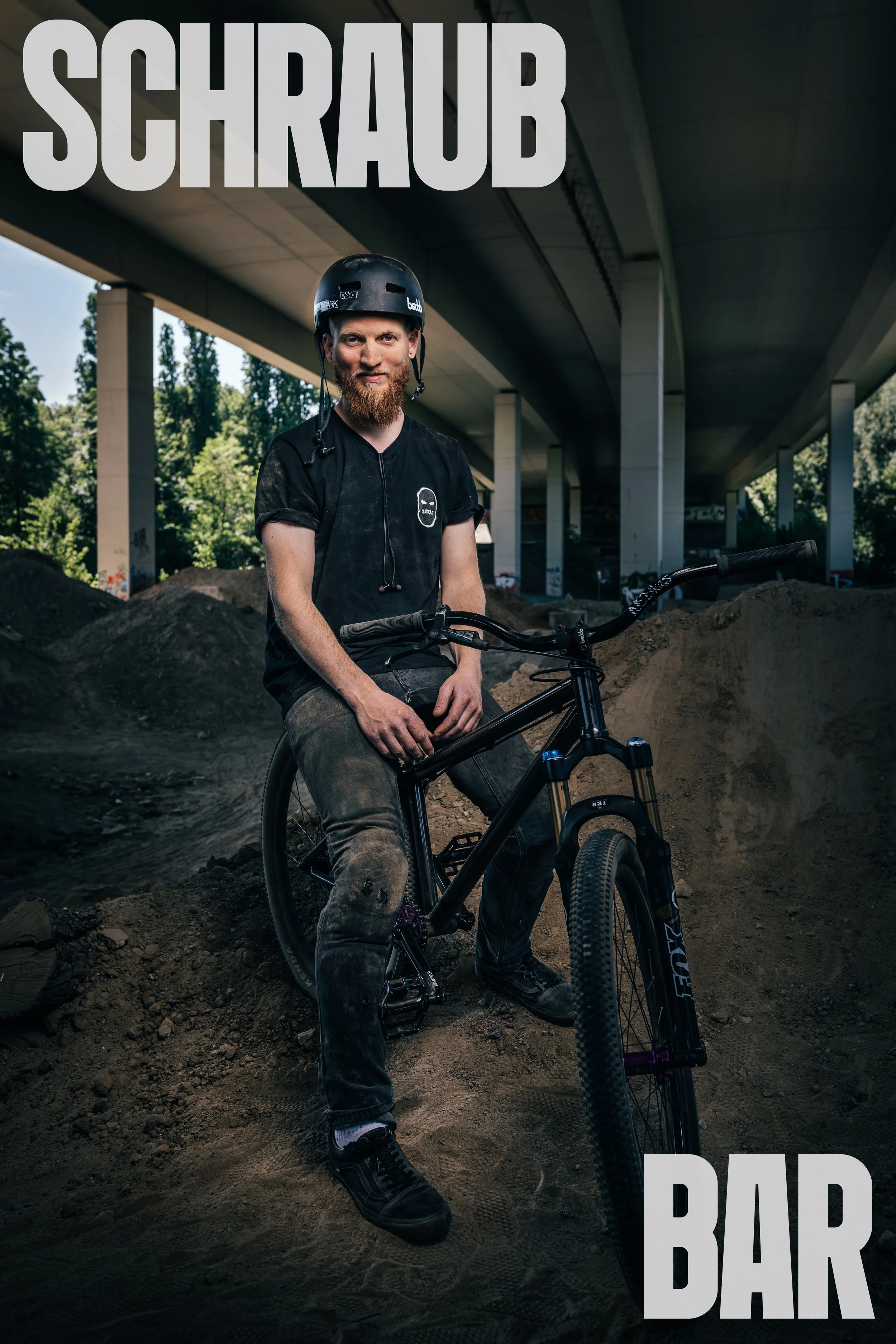Ein junger Mann mit Fahrrad unter einer Brücke, trägt Helm und schwarze T-Shirt, umgeben von Sand und Erdhügel, Text 'Schraub Bar' im oberen und unteren Bereich.