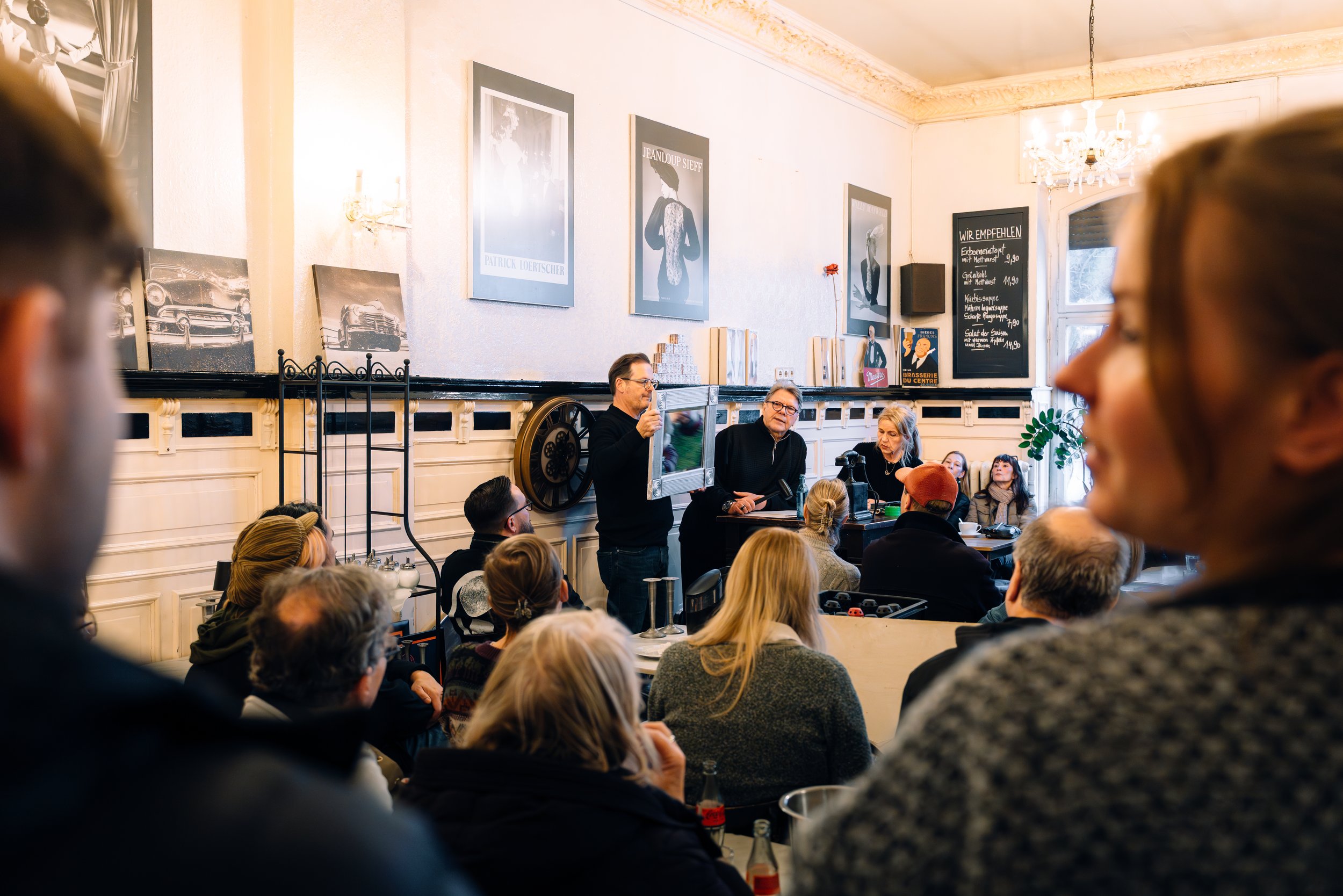 Ein Mann hält ein Gemälde hoch, während er auf einer kleinen Bühne spricht. Es sitzen mehrere Menschen im Raum, die einem Vortrag oder einer Lesung zuhören. Der Raum ist elegant mit Bildern, einem Kronleuchter und einer schwarzen Tafel mit Speisekart