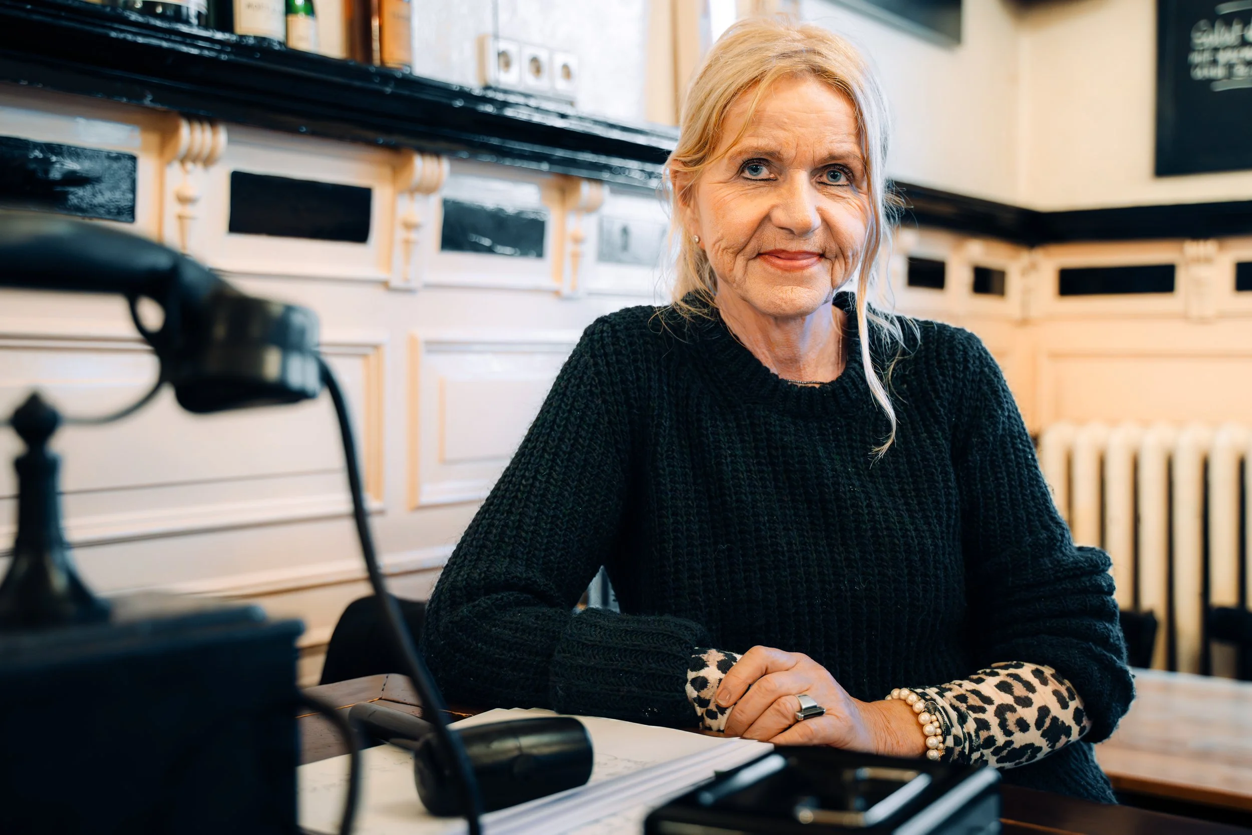 Eine ältere Frau sitzt in einem Café, trägt einen schwarzen Pullover mit Leopardenmuster an den Ärmeln und hat blonde Haare. Sie sitzt an einem Tisch mit einem Notizbuch, einem Stift und einem Telefon. Im Hintergrund sind Regale mit Büchern und eine 