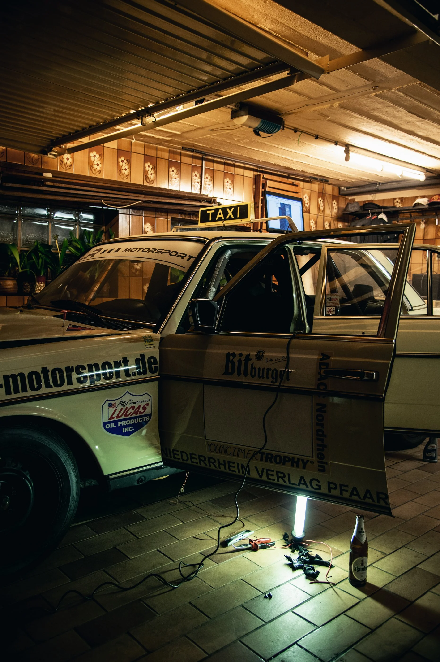Ein Motorsportwagen im Keller, mit geöffneten Autotüren, davor ein Arbeitslicht, Werkzeuge und eine Flasche Bier auf dem Boden, an der Wand sind Pflanzen und ein Bildschirm.