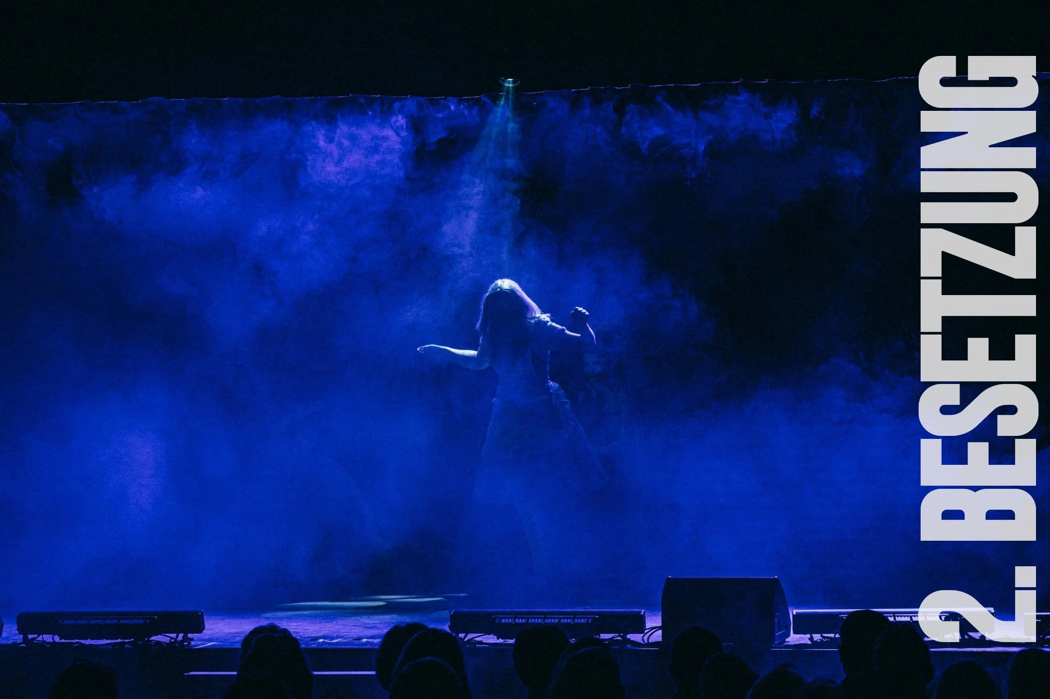 Eine Person tanzt auf einer dunklen Bühne, umgeben von blauer Nebel, mit der Aufschrift 2.BEESZEUZNG auf der rechten Seite.