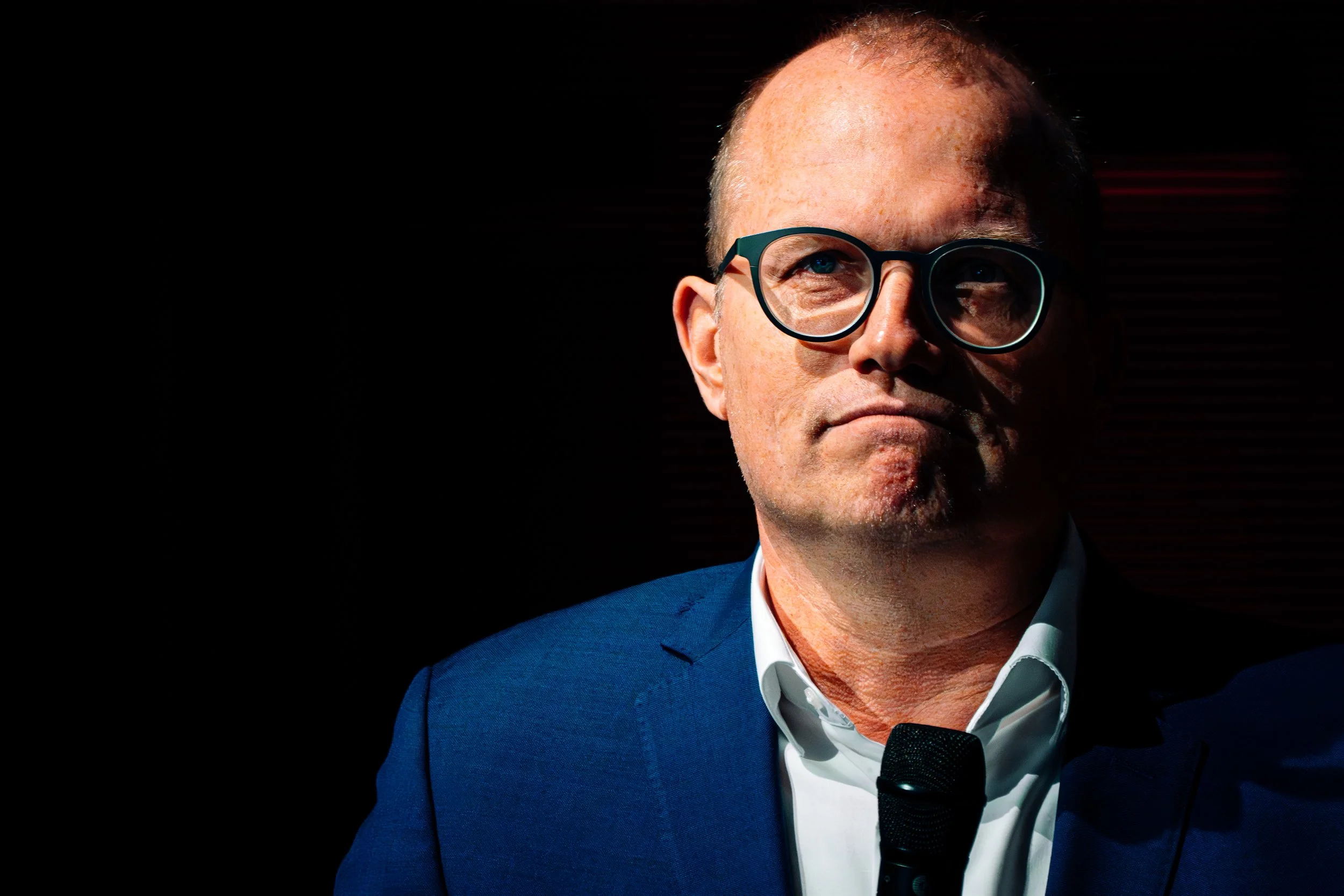 Ein Mann in einem blauen Anzug und weißen Hemd mit schwarzen Brillen hält ein Mikrofon, dunkler Hintergrund.