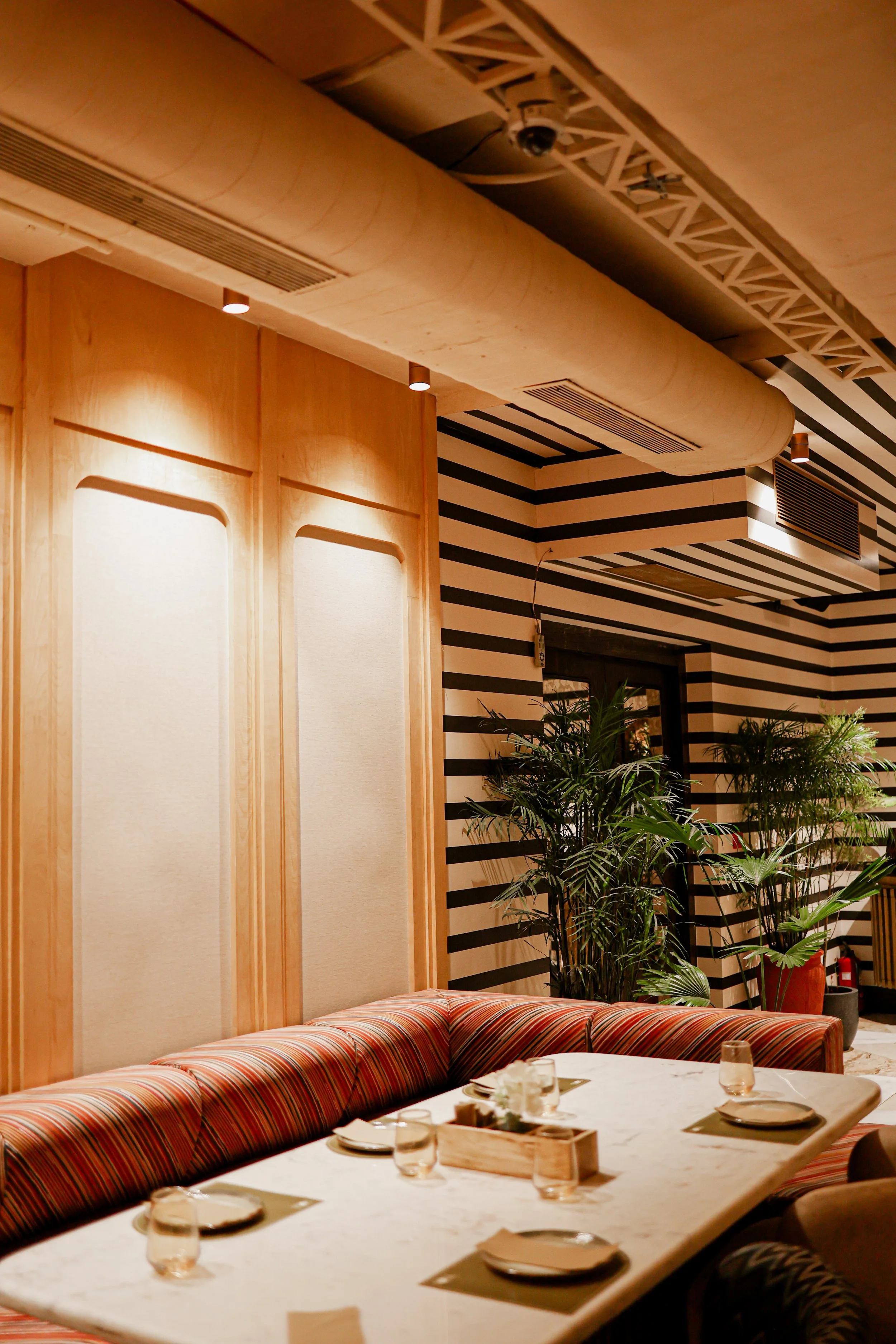 Interior of a restaurant with a marble table set with plates, glasses, and napkins, surrounded by a striped upholstered banquette, wood-paneled walls, and potted plants, with black-and-white striped walls and ceiling features.