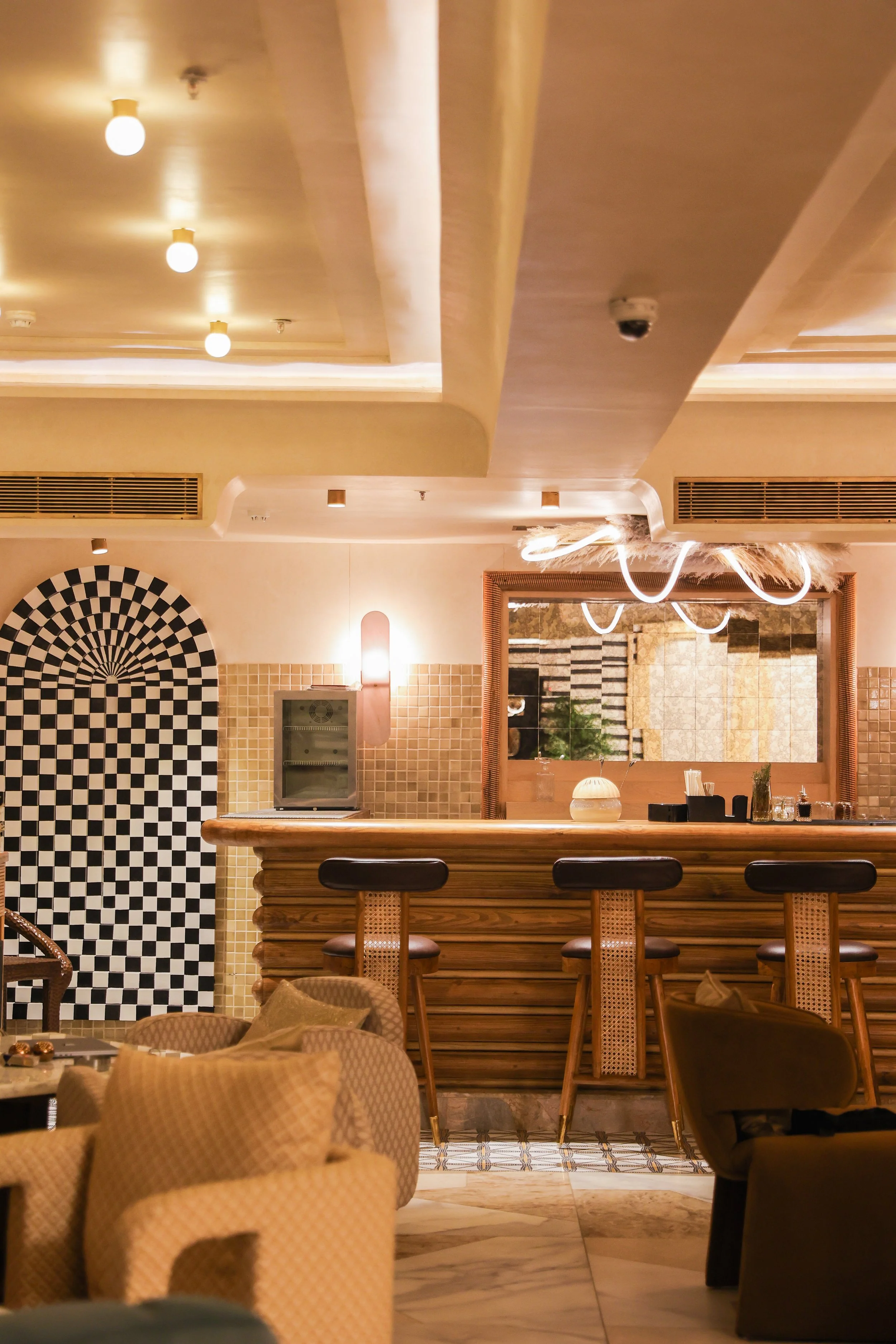 Interior of a stylish bar with a wooden counter, black bar stools, patterned seating, and modern lighting features.