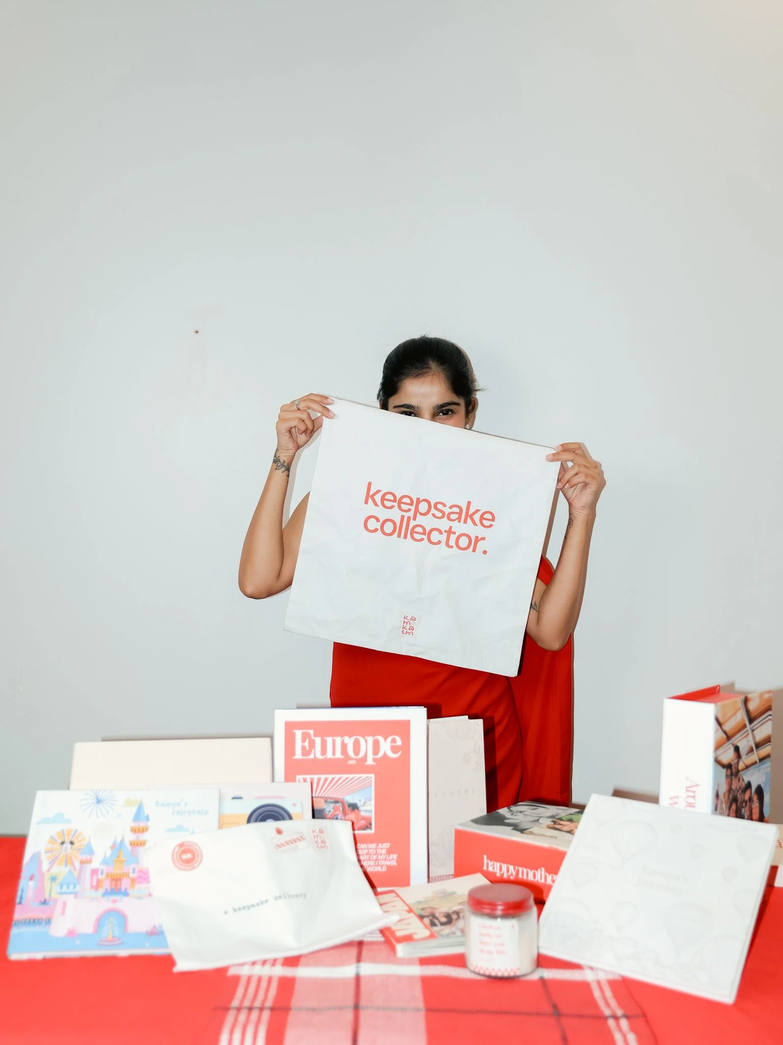 A woman in a red shirt holding a white bag with red text that reads 'keepsake collector'. She stands behind a table covered with various colorful books and boxes on a red checkered tablecloth.