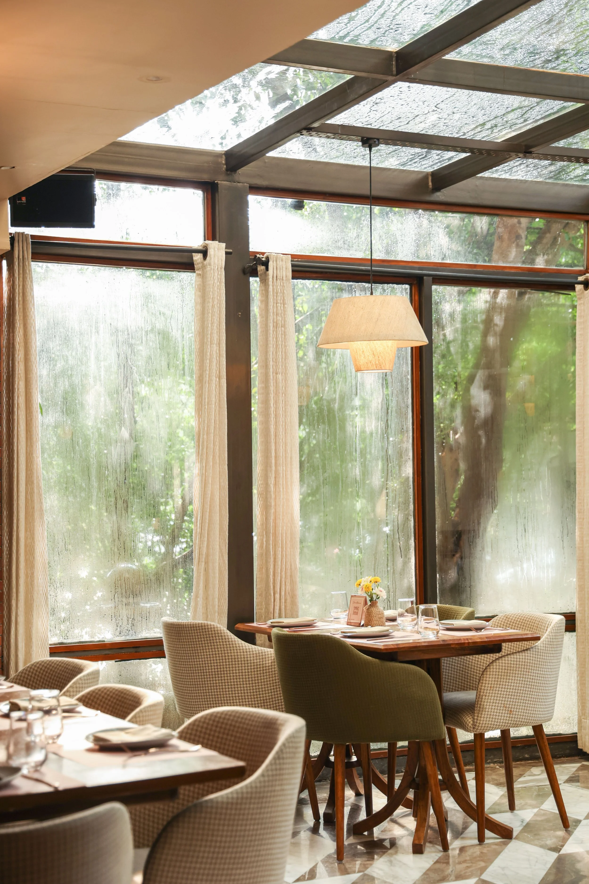 A cozy restaurant interior with large windows, beige curtains, a hanging lamp, and a wooden table set with glassware, plates, and a small flower arrangement.