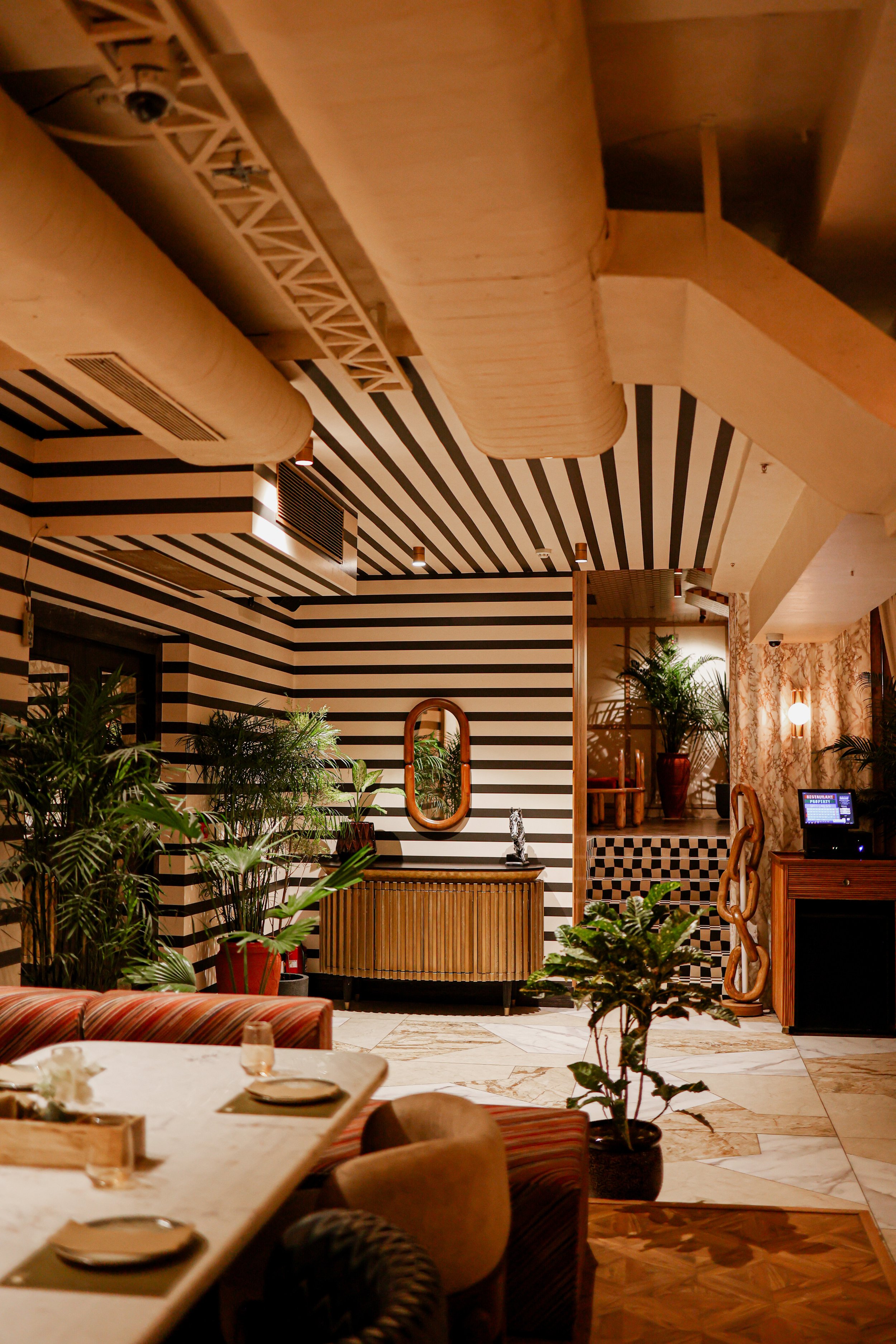 Interior of a restaurant or hotel with striped black and white walls, lush green plants, wooden furniture, and decorative mirrors and chains, creating a stylish and cozy atmosphere.