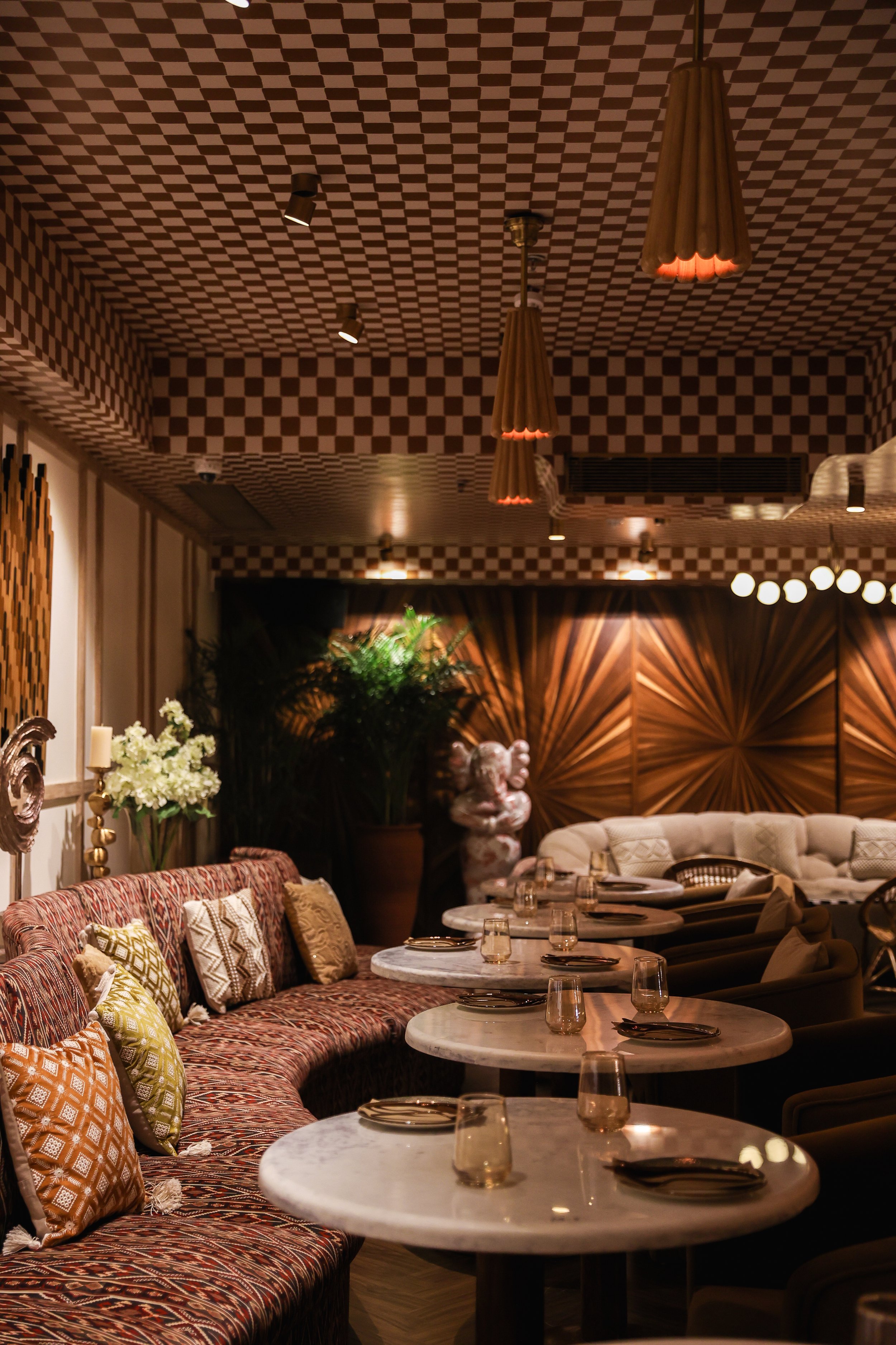 Interior of a restaurant with marble-top tables set with plates, glasses, and cutlery, a patterned curved bench with decorative pillows, plush chairs, and warm lighting fixtures.