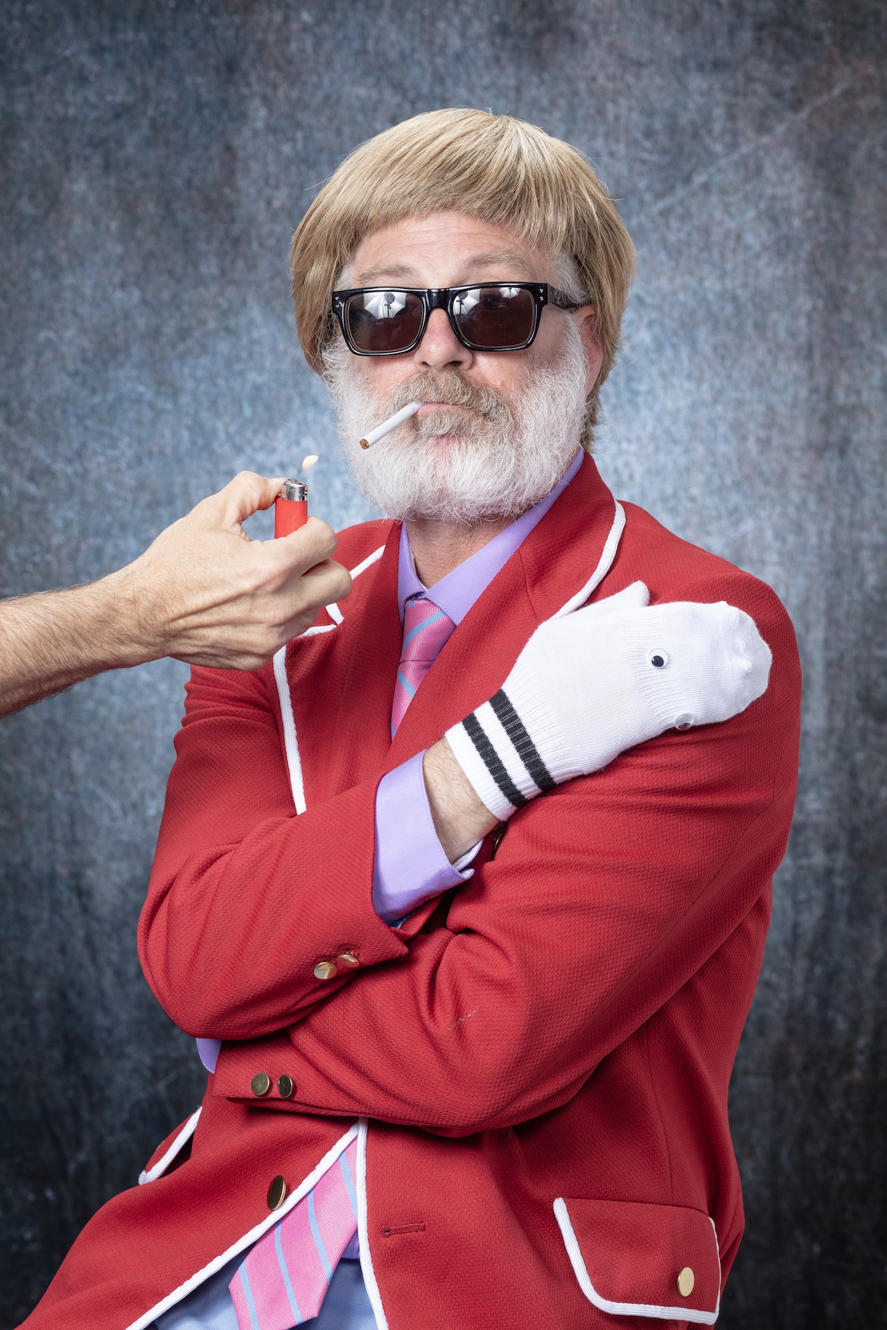 A man with gray hair and a beard wearing black sunglasses and a red blazer with white piping, holding a white glove and being attended to with a cigarette in his mouth and a lighter in hand.