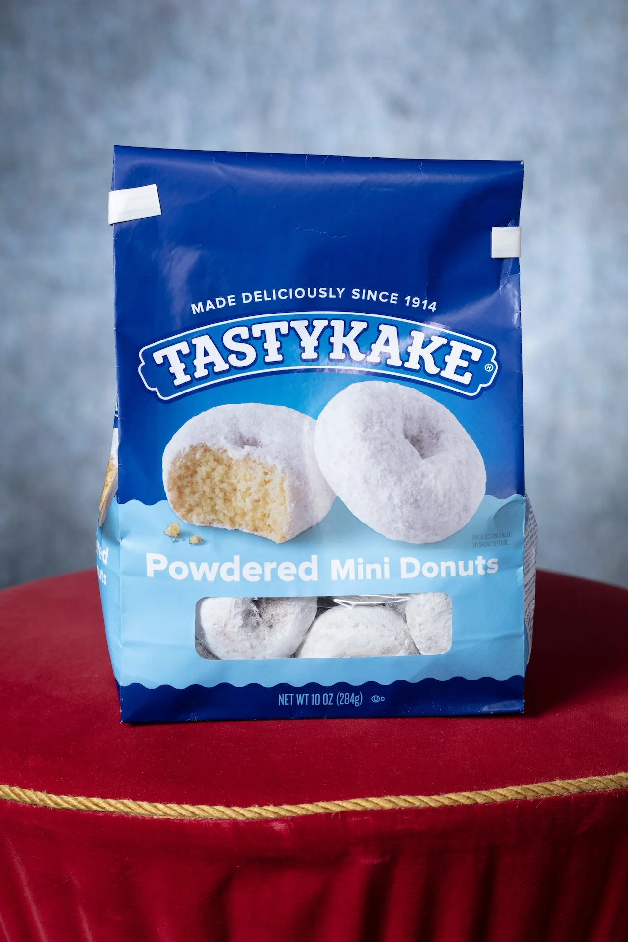 A blue bag of Tastykake Powdered Mini Donuts, with a window showing powdered mini donuts inside, placed on a red velvet surface.
