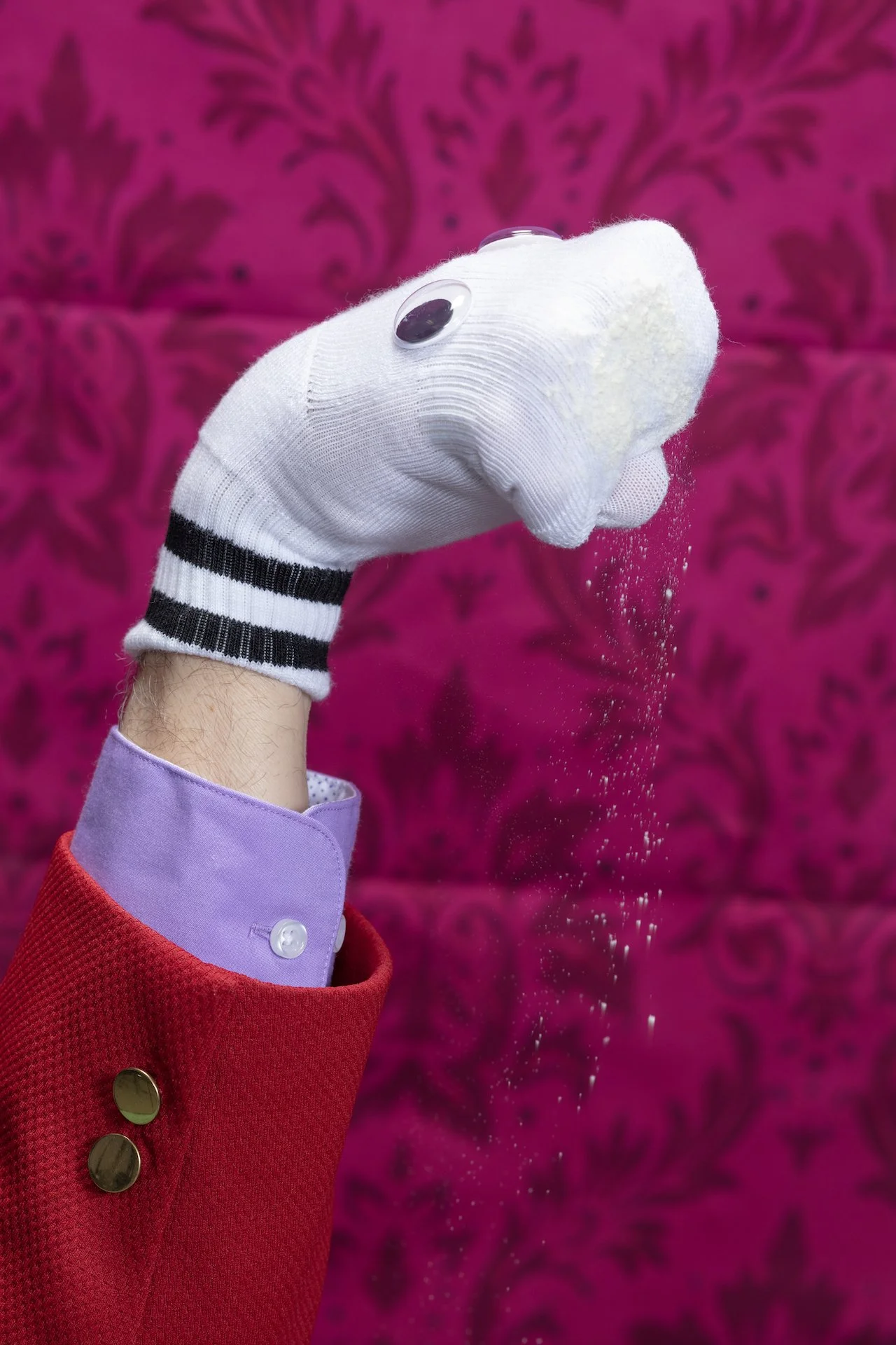 A puppet or toy dog with googly eyes, wearing a sock as a nose, being held by a person in a red coat. The person has a purple shirt with a white button visible at the cuff.