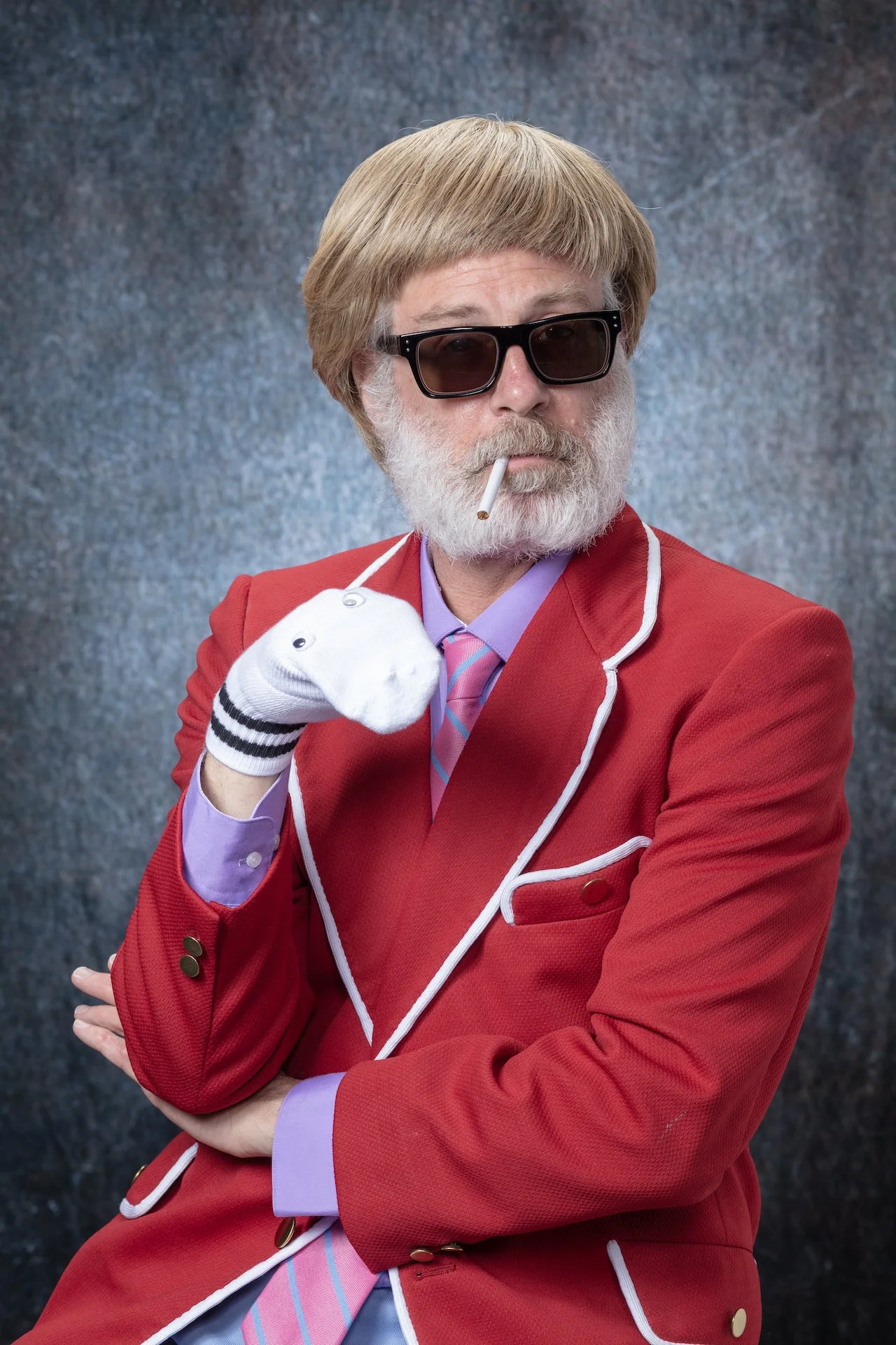 A man with blonde hair, beard, and sunglasses wearing a red blazer, purple shirt, and striped tie, posing with a stuffed sock puppet nose in his shirt pocket against a grey textured background.