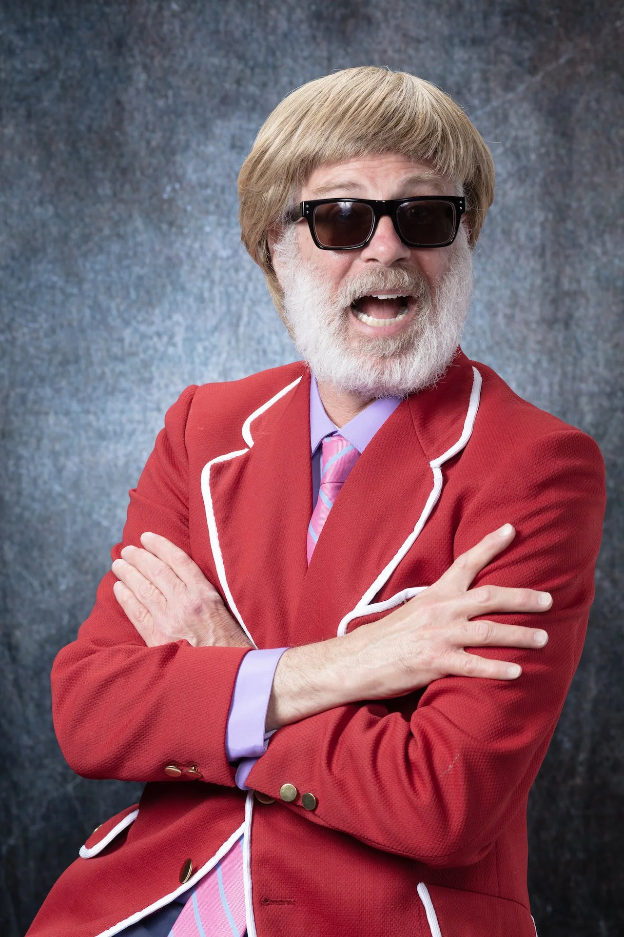 A man with a gray beard and hair, wearing large black sunglasses, a red blazer with white piping, a pink and blue striped tie, and a light purple dress shirt, standing against a dark textured background.