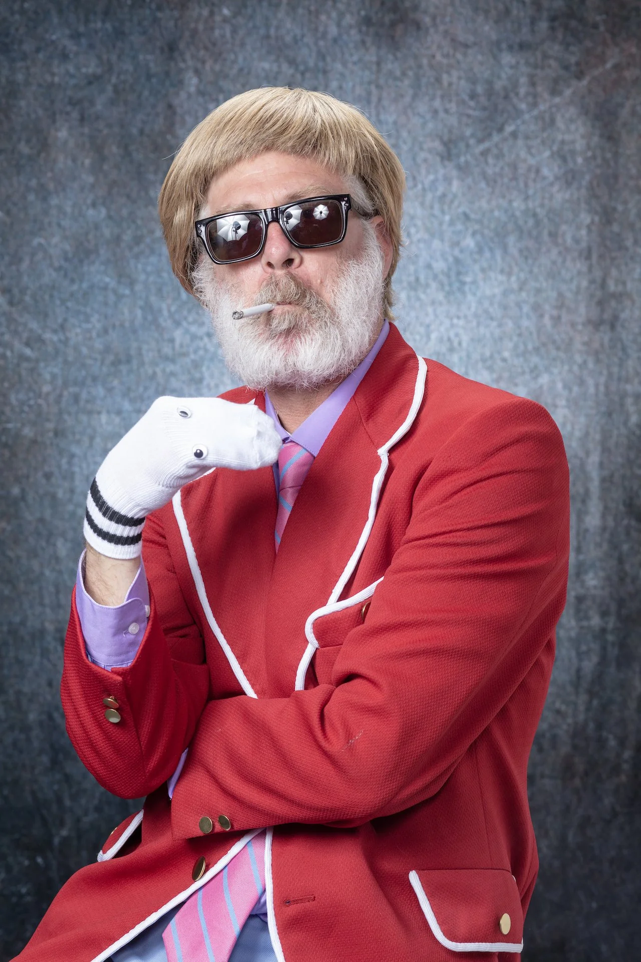 An elderly man with a white beard and blond hair wearing dark sunglasses, a red blazer with white trim, a purple shirt, and a pink striped tie. He has a cigarette in his mouth and is gesturing with his right hand near his chin, as if smoking or posin